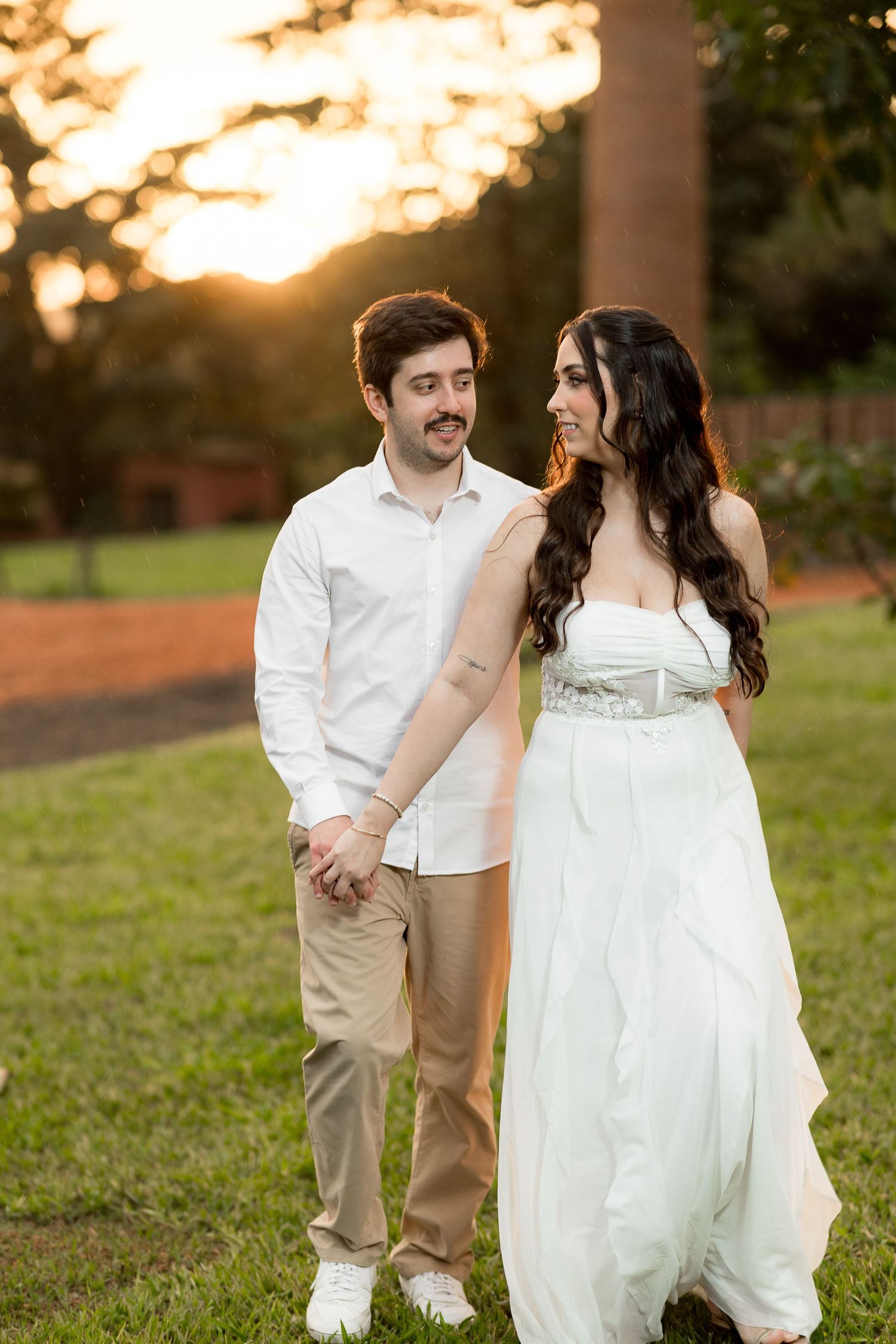 Couple holding hands at sunset in a park, woman in white strapless gown, man in white shirt and khakis.