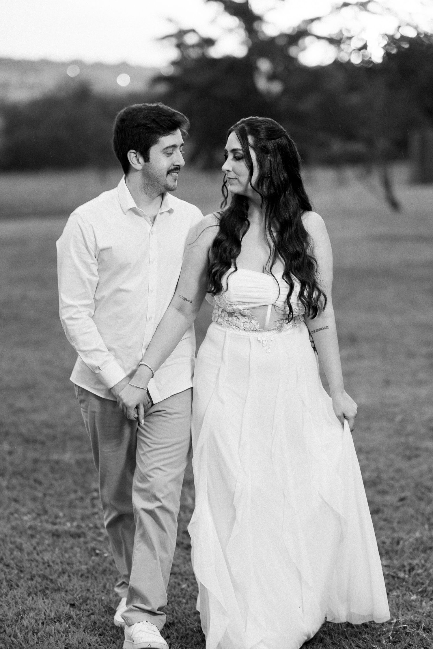 Black and white photo of a couple in white attire holding hands and smiling at each other in an outdoor setting.