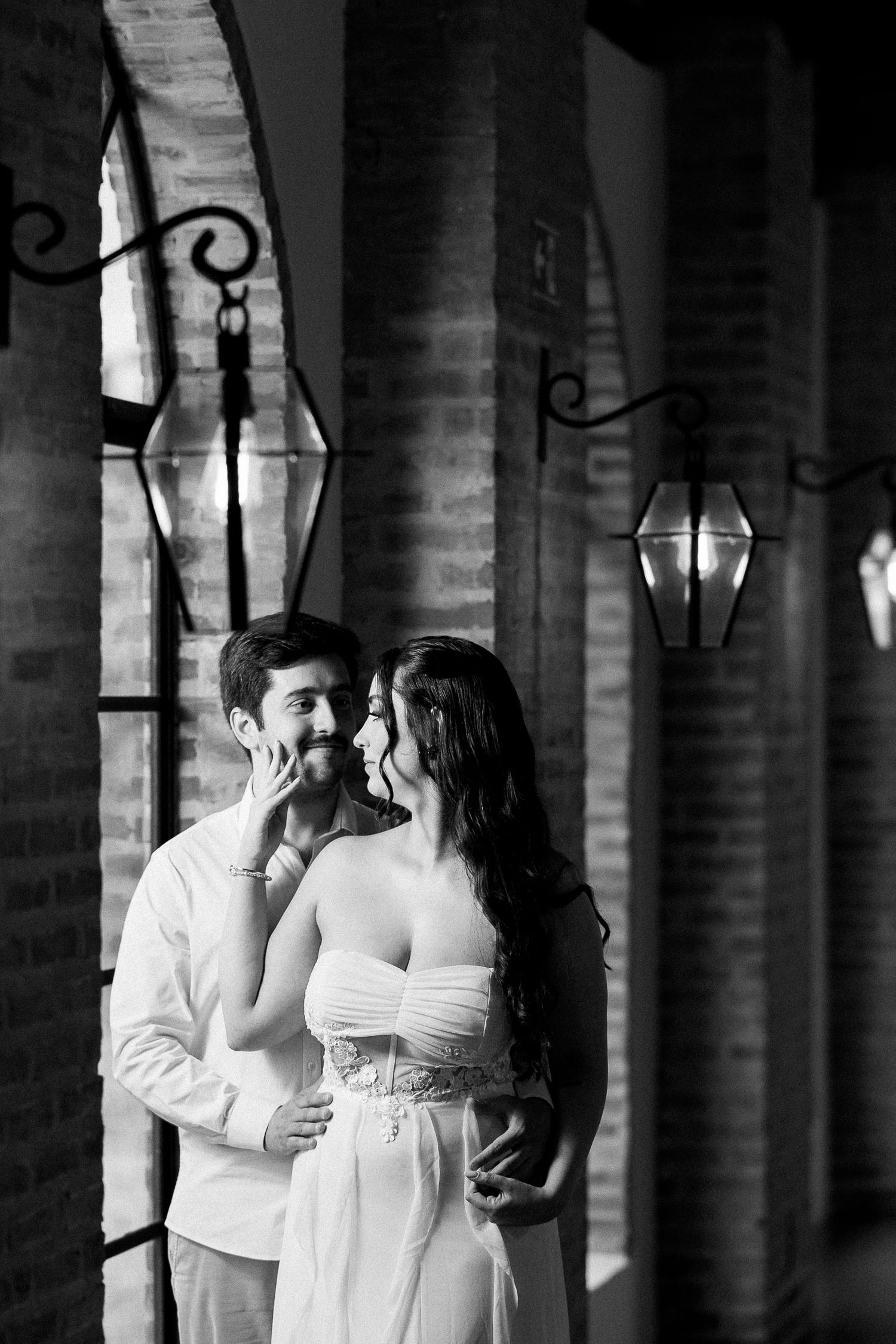 Black and white photo of a romantic couple embracing near brick arches with vintage lanterns.