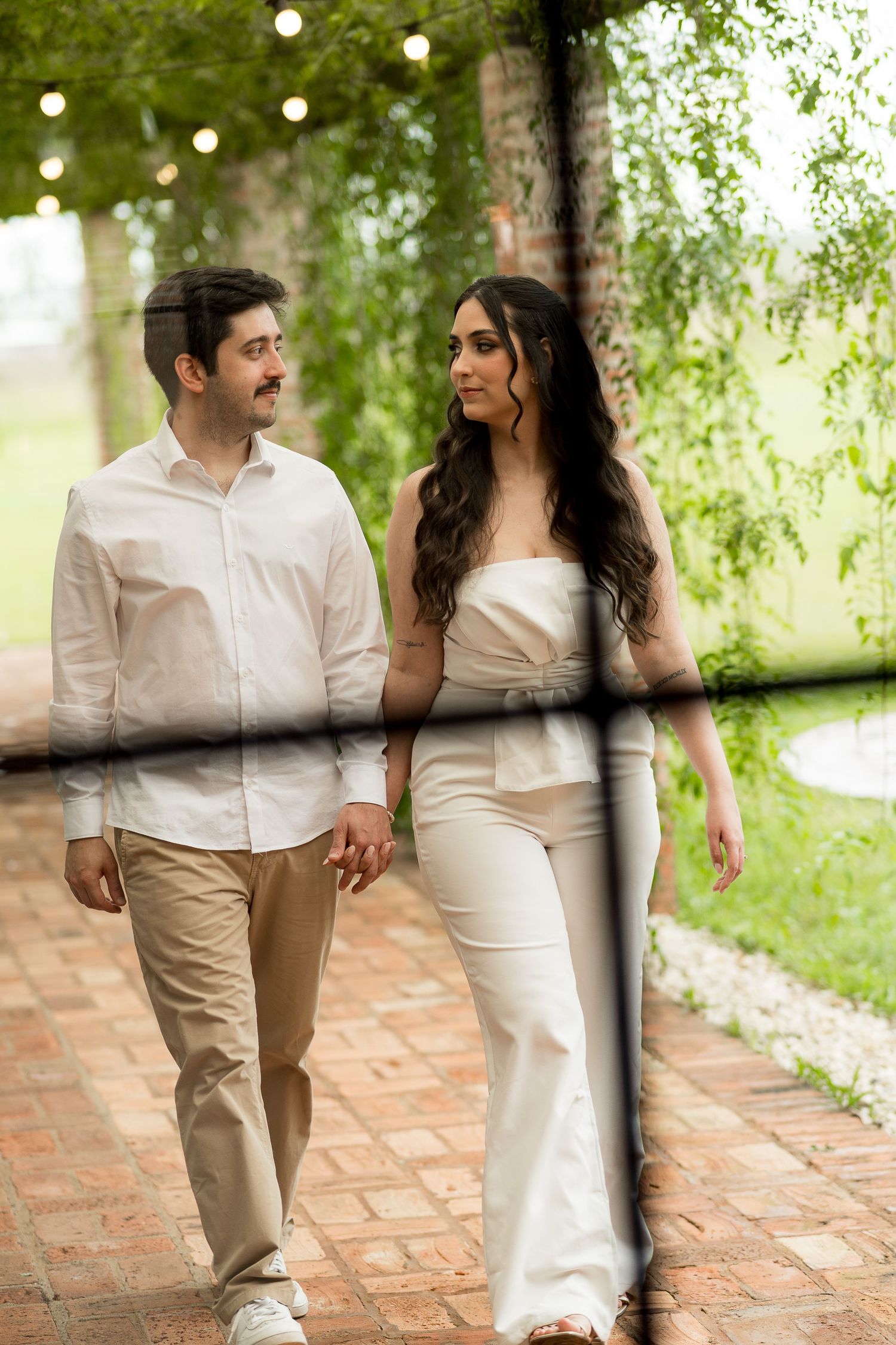 Couple in white outfits walking hand in hand through a lush garden pathway with string lights overhead.