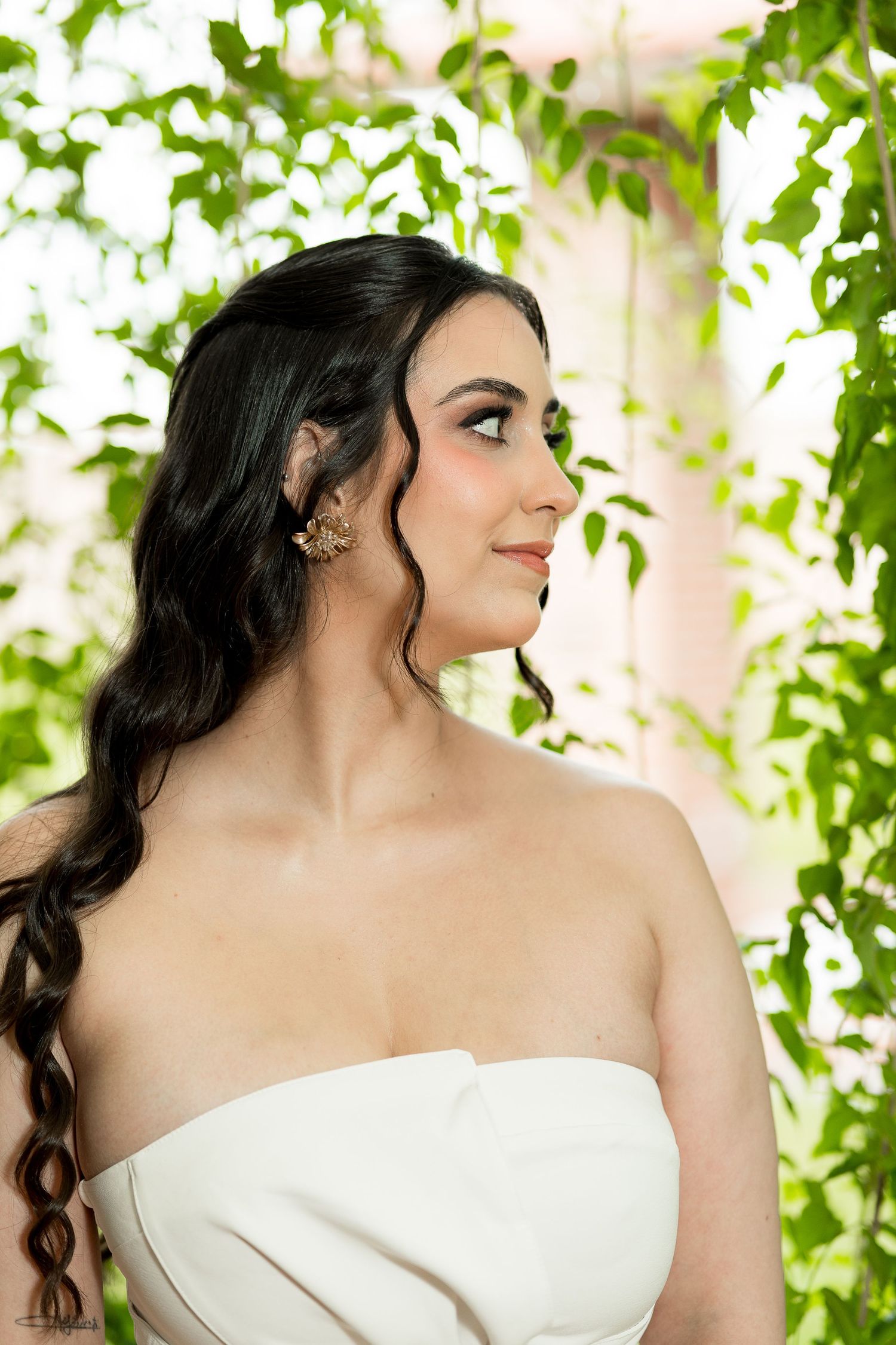 Elegant bride in strapless white gown with gold earrings, dark curly hair, surrounded by lush green foliage.