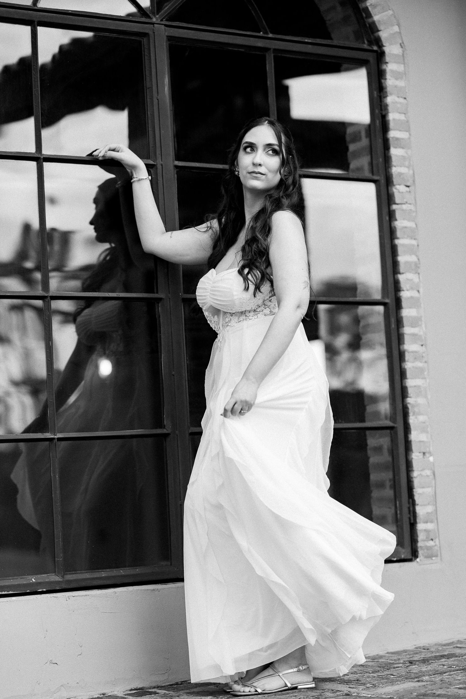 Black and white photo of a woman in a flowing white gown posing elegantly by a large arched window.