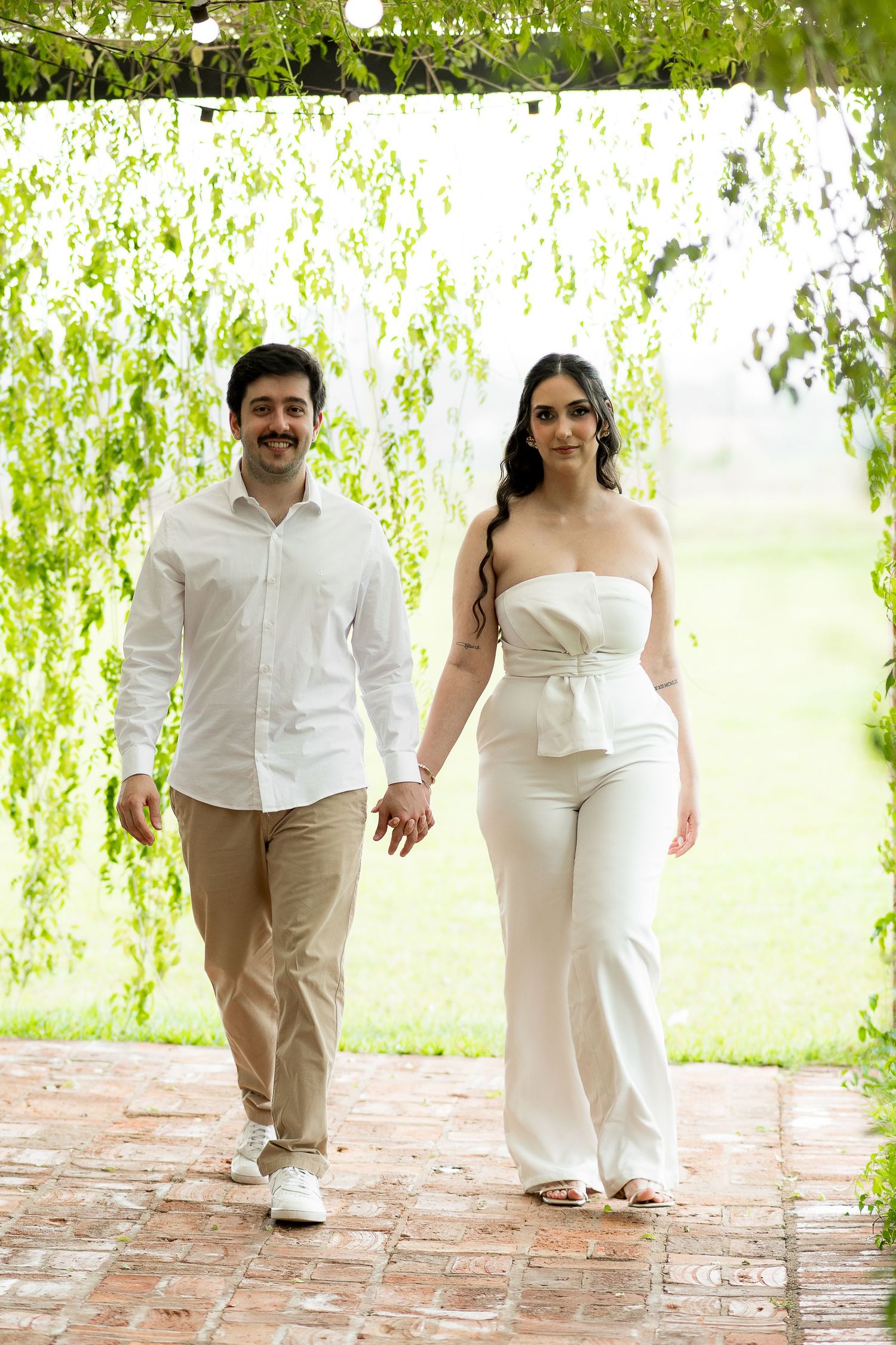 A smiling couple in white outfits walks hand in hand on a brick path surrounded by lush green weeping willow trees.