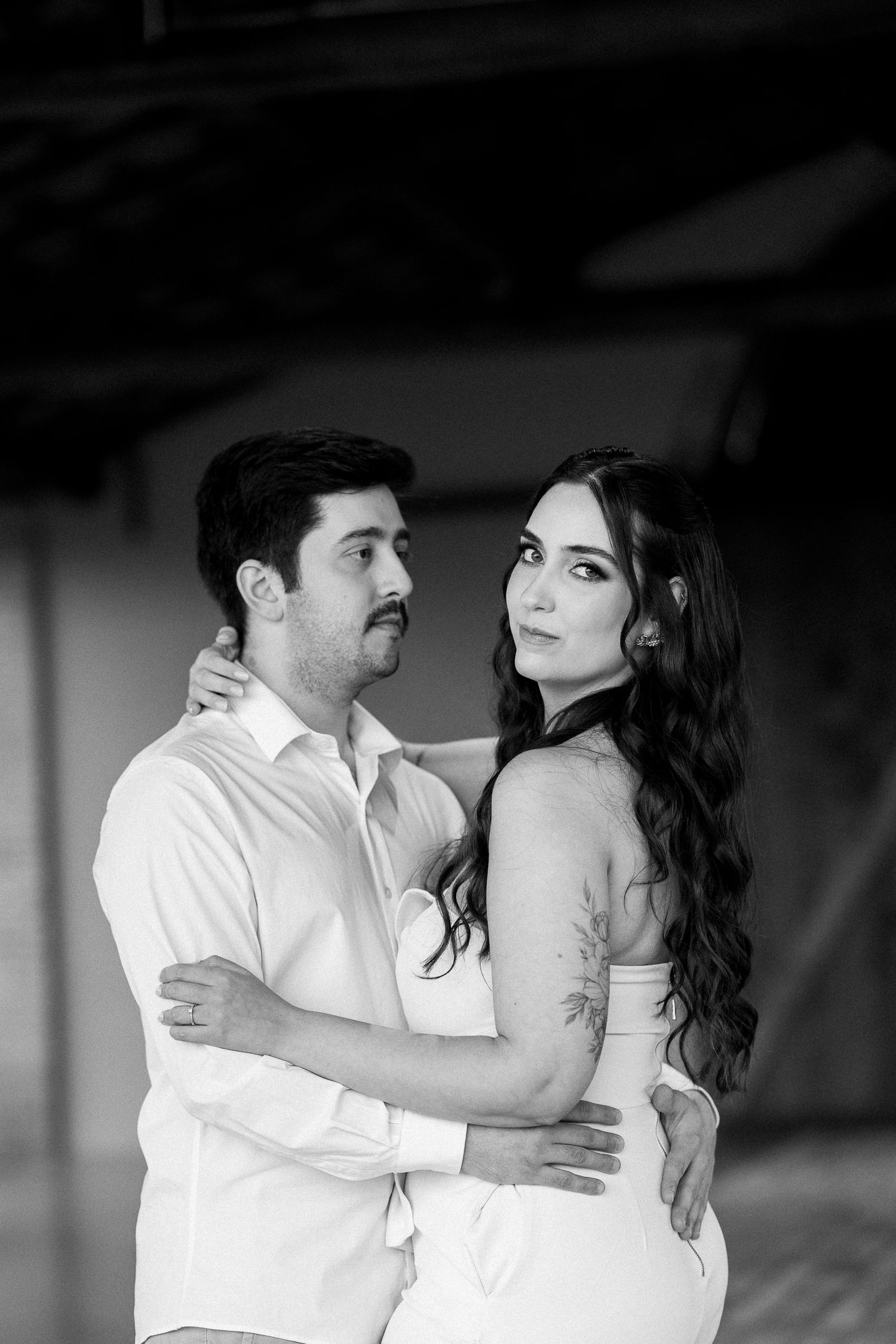 Black and white photo of a couple embracing, woman with long wavy hair and tattoos in white dress, man in white shirt.