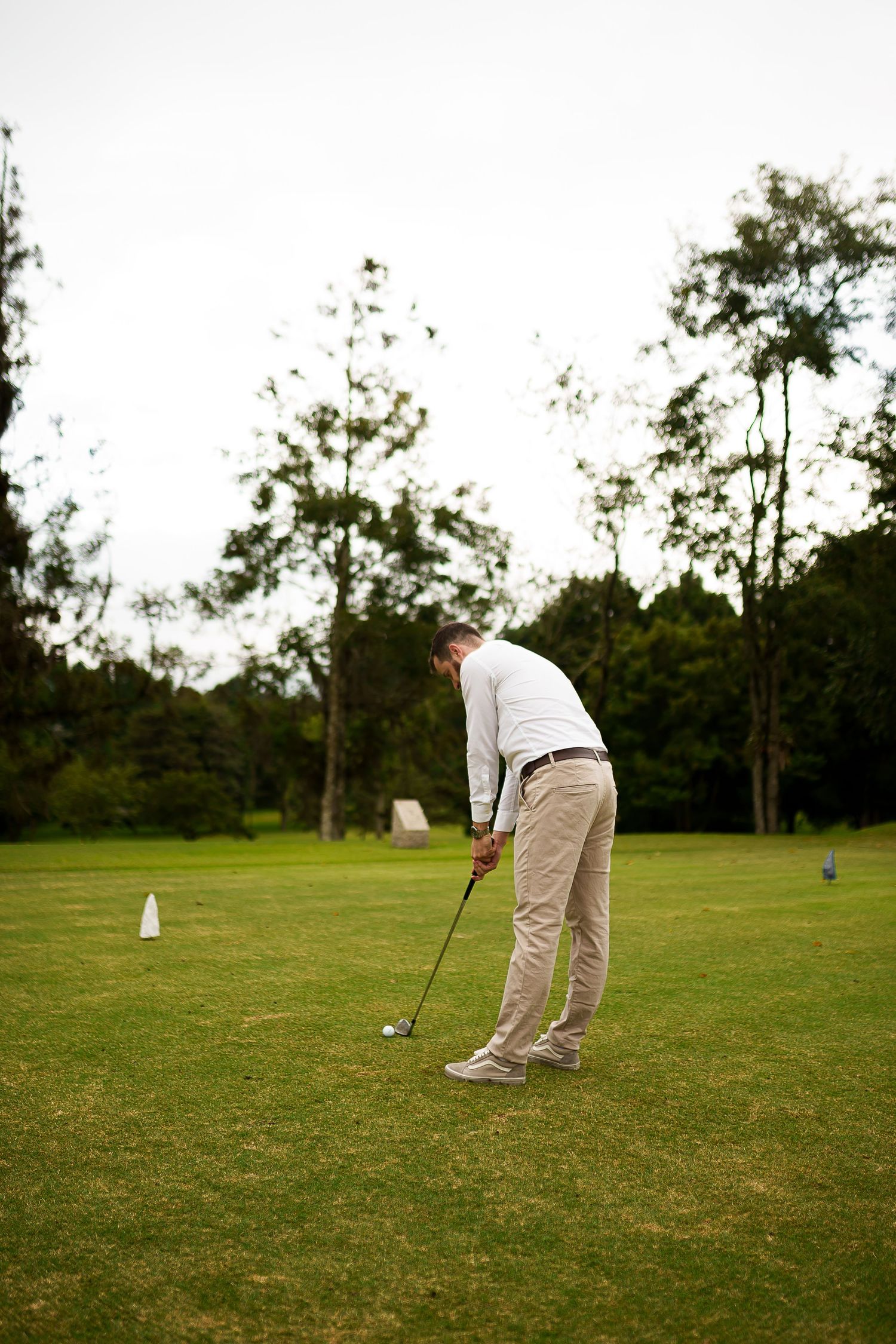 A golfer in white shirt and khaki pants takes a swing on a lush green golf course surrounded by trees.