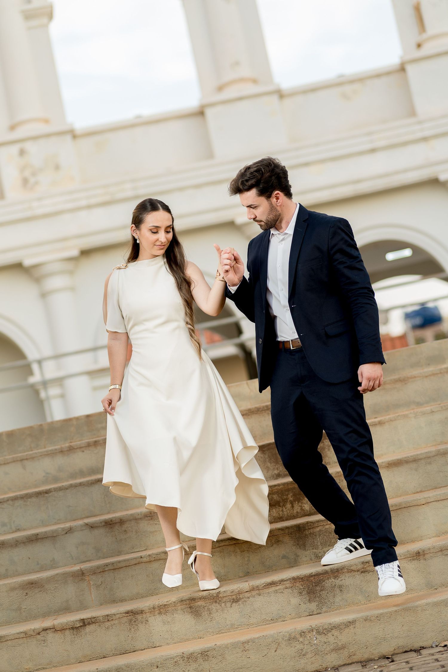 A couple in formal attire walks down stone steps, man in suit kisses womans hand while she wears elegant white midi dress.
