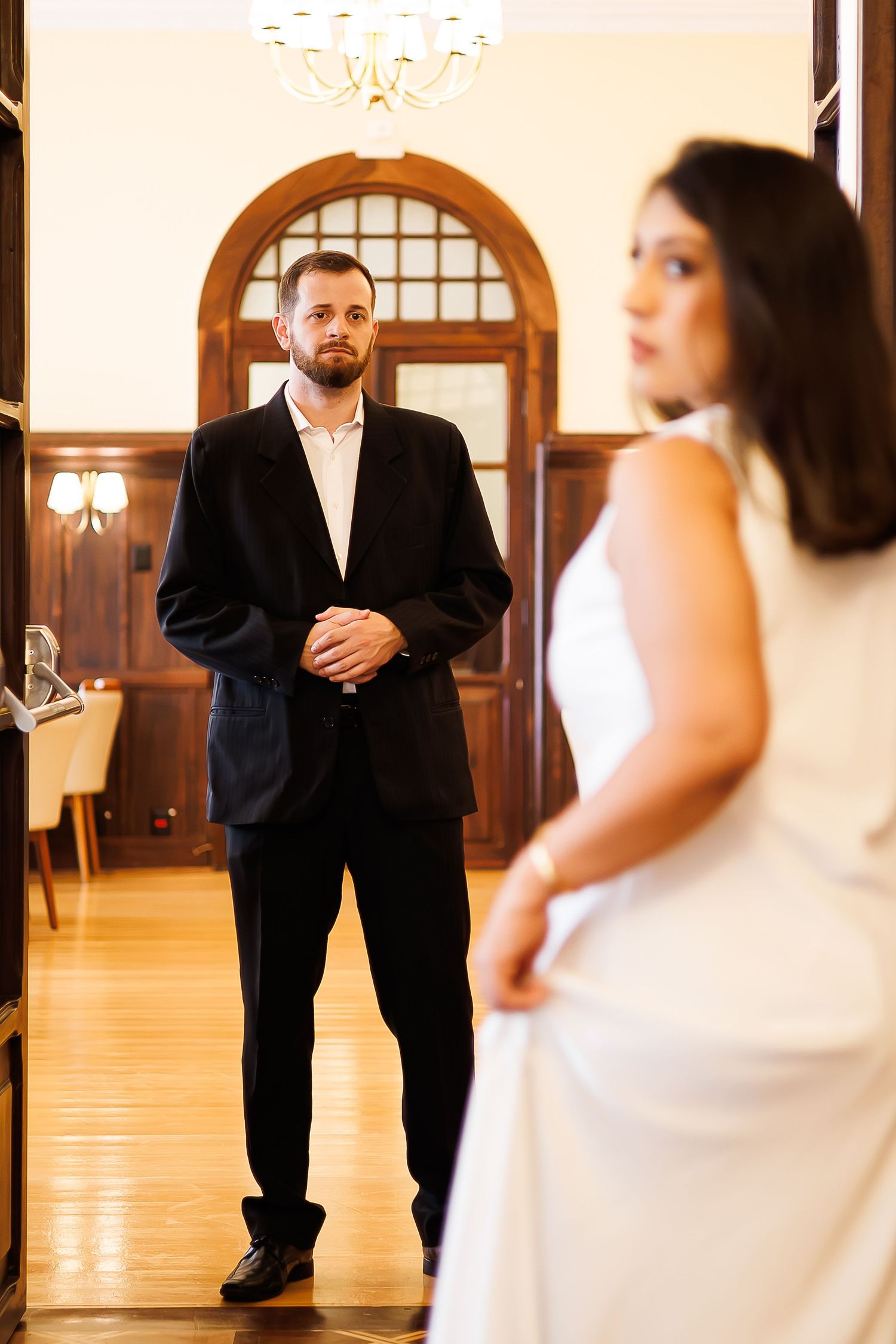 Groom in black suit gazes at bride in white dress standing in elegant wood-paneled room with chandelier.