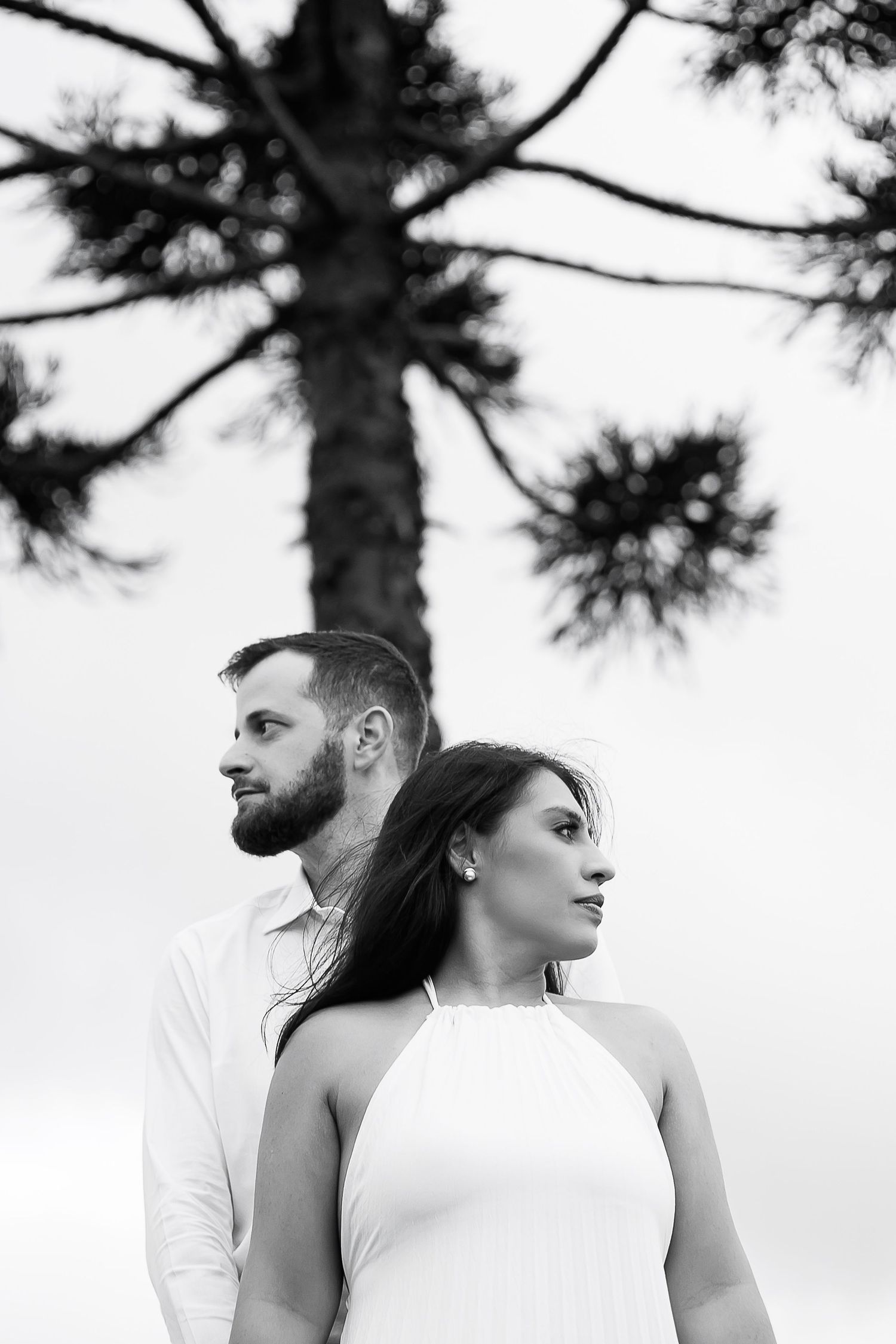 Black and white photo of a couple standing back to back under a pine tree, gazing into the distance in white outfits.