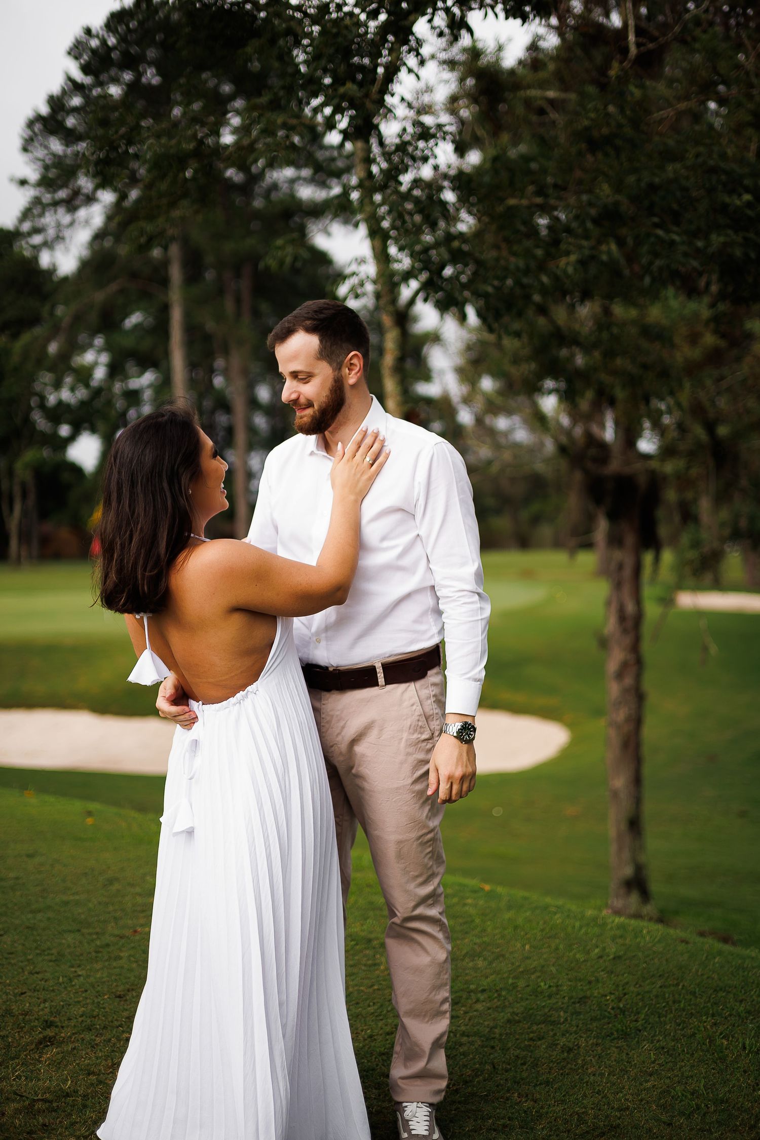 Couple in white attire sharing a romantic moment on a lush golf course surrounded by tall trees.