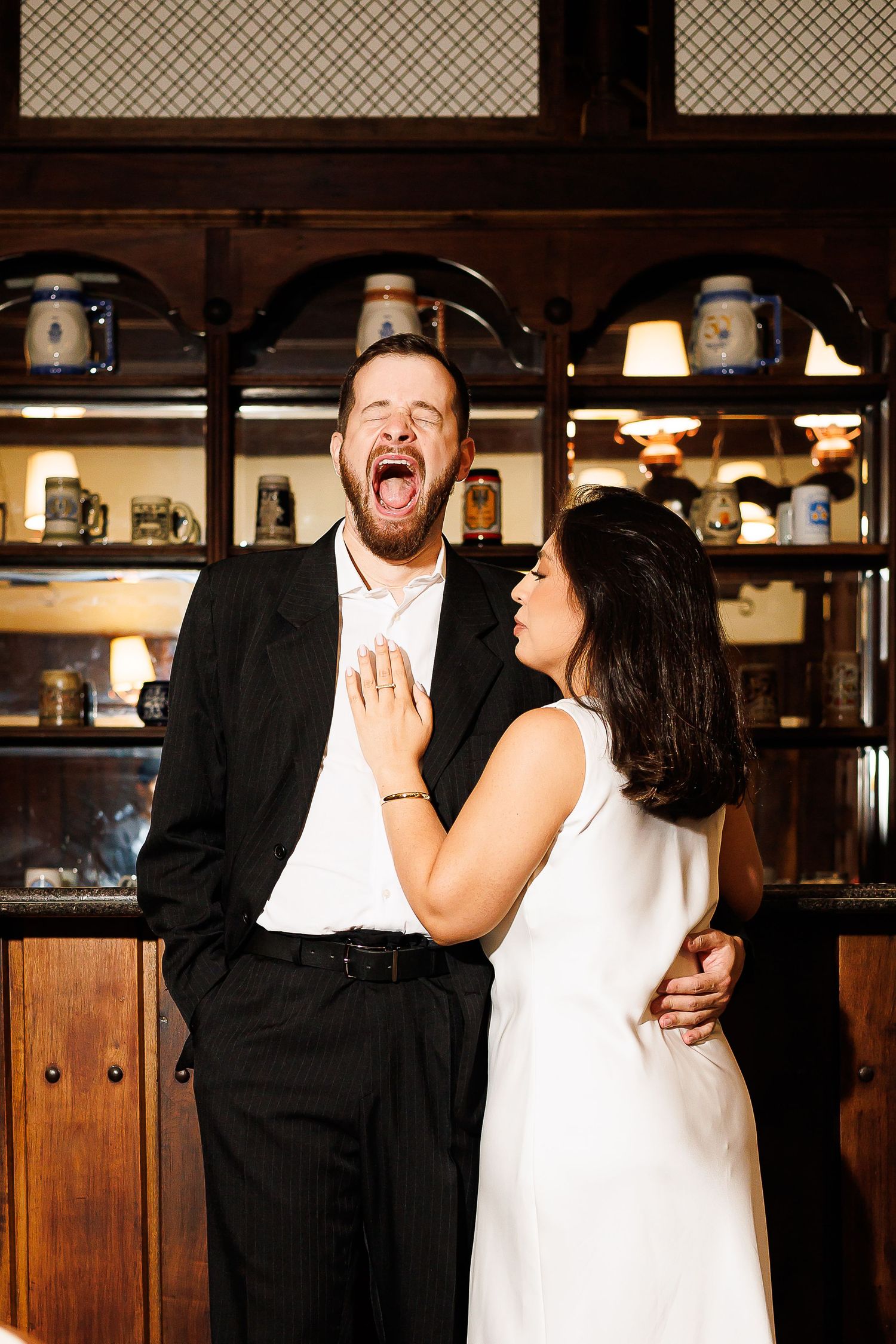 Laughing couple in formal wear posing together in front of wooden shelves displaying beer steins.