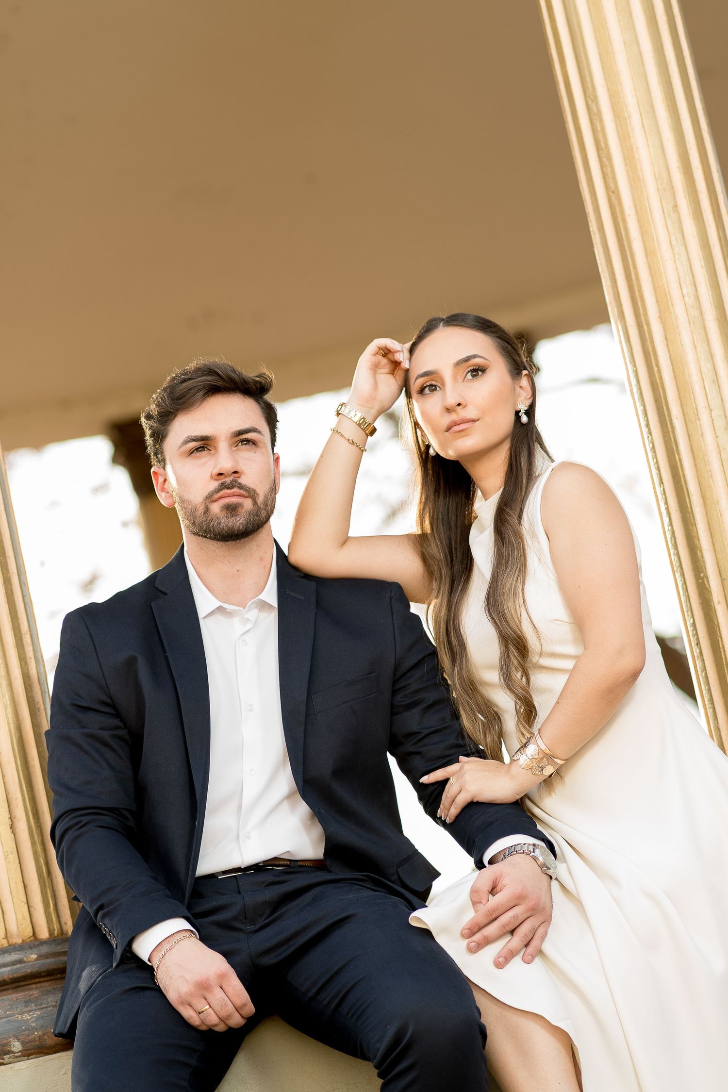 Elegant couple in formal attire sitting between golden columns, woman in white dress touching hair, man in black suit.