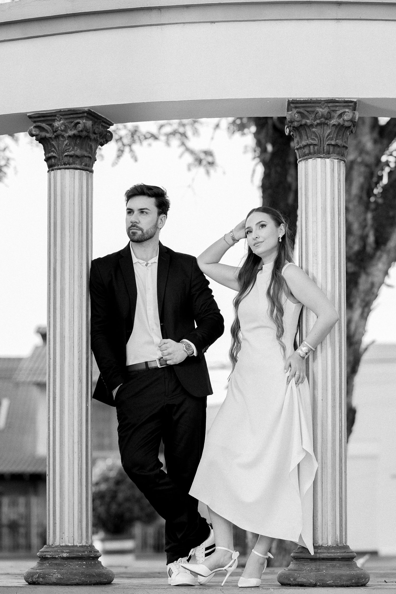 Black and white photo of elegant couple posing between classical columns, man in suit, woman in white dress.