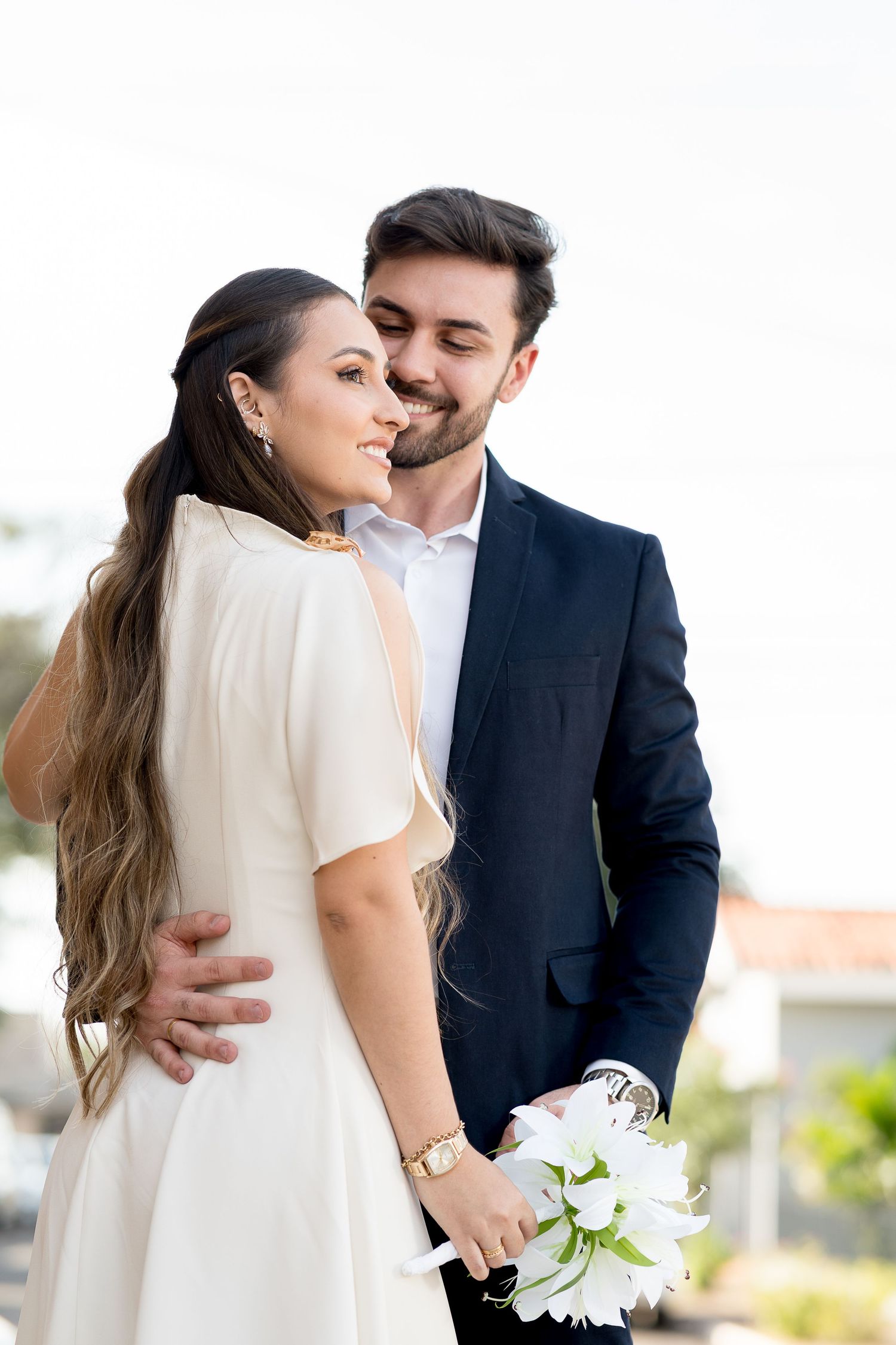 Couple in wedding attire embracing outdoors, bride in white dress holding bouquet, groom in navy suit smiling.