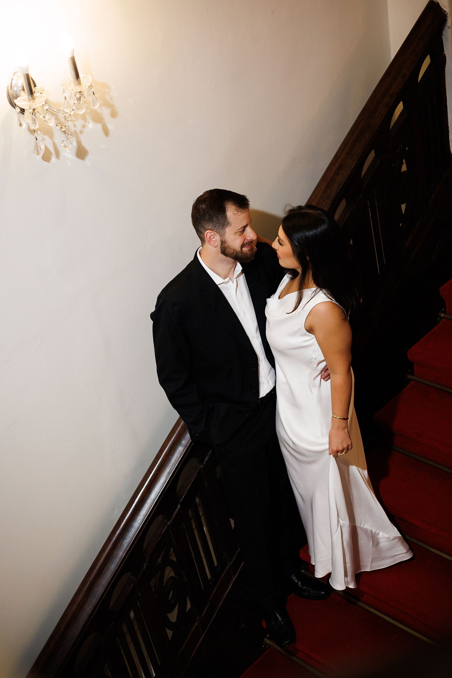 Bride and groom share a romantic moment on a grand staircase, she in white gown, he in black tuxedo.