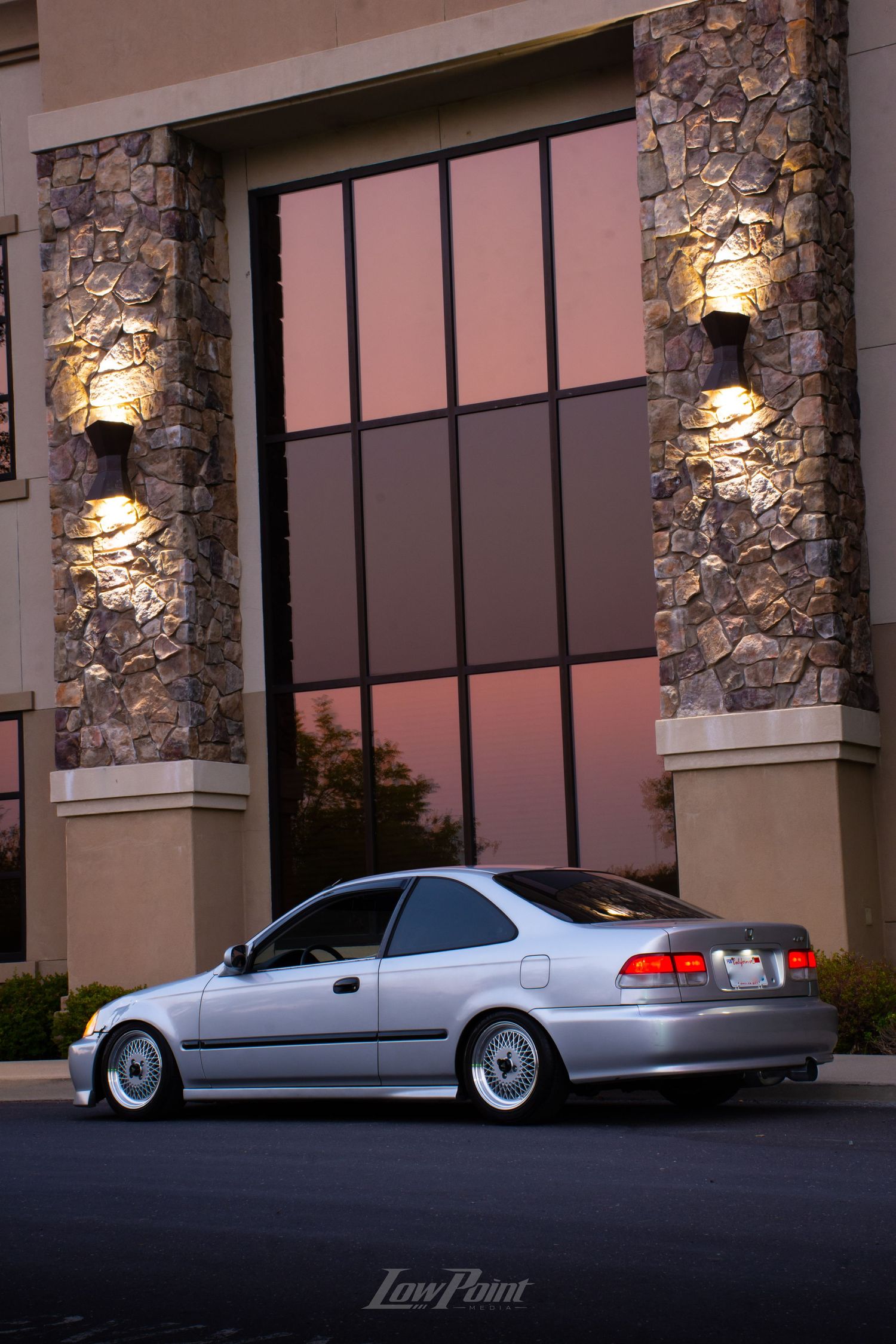 Silver Honda Civic photographed in front of modern building with large windows reflecting a colorful pink sunset