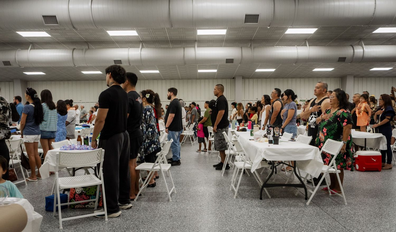 Crowd of people mingling at indoor cultural event with white folding chairs and tables in large hall.