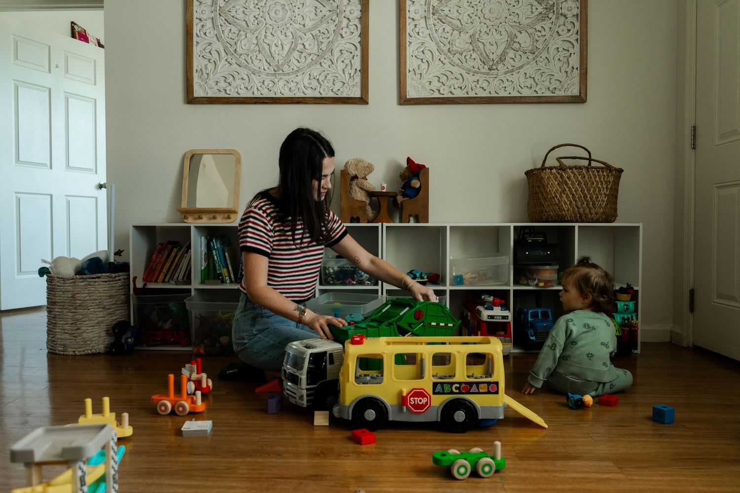 Mother sitting on floor playing with toddler and yellow school bus toy in cozy living room.