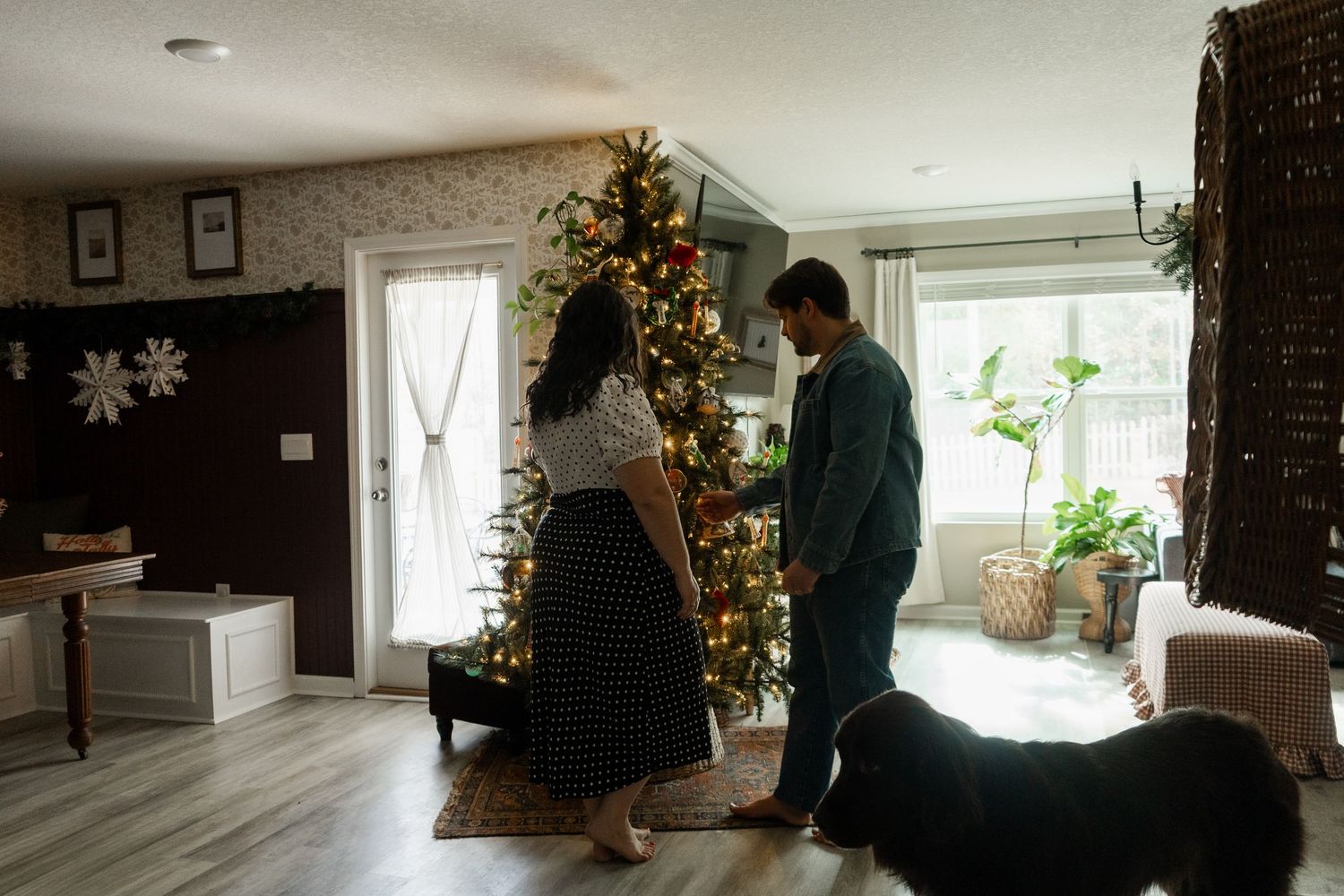 Couple standing near decorated Christmas tree with dog in cozy living room holiday home decor.