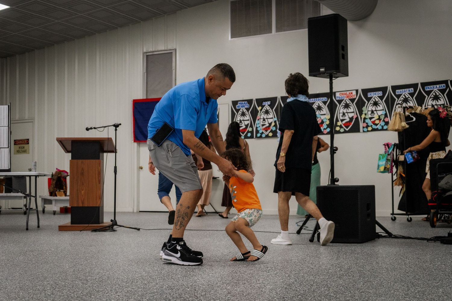Man in blue shirt dances playfully with young child in orange shirt at indoor community event.