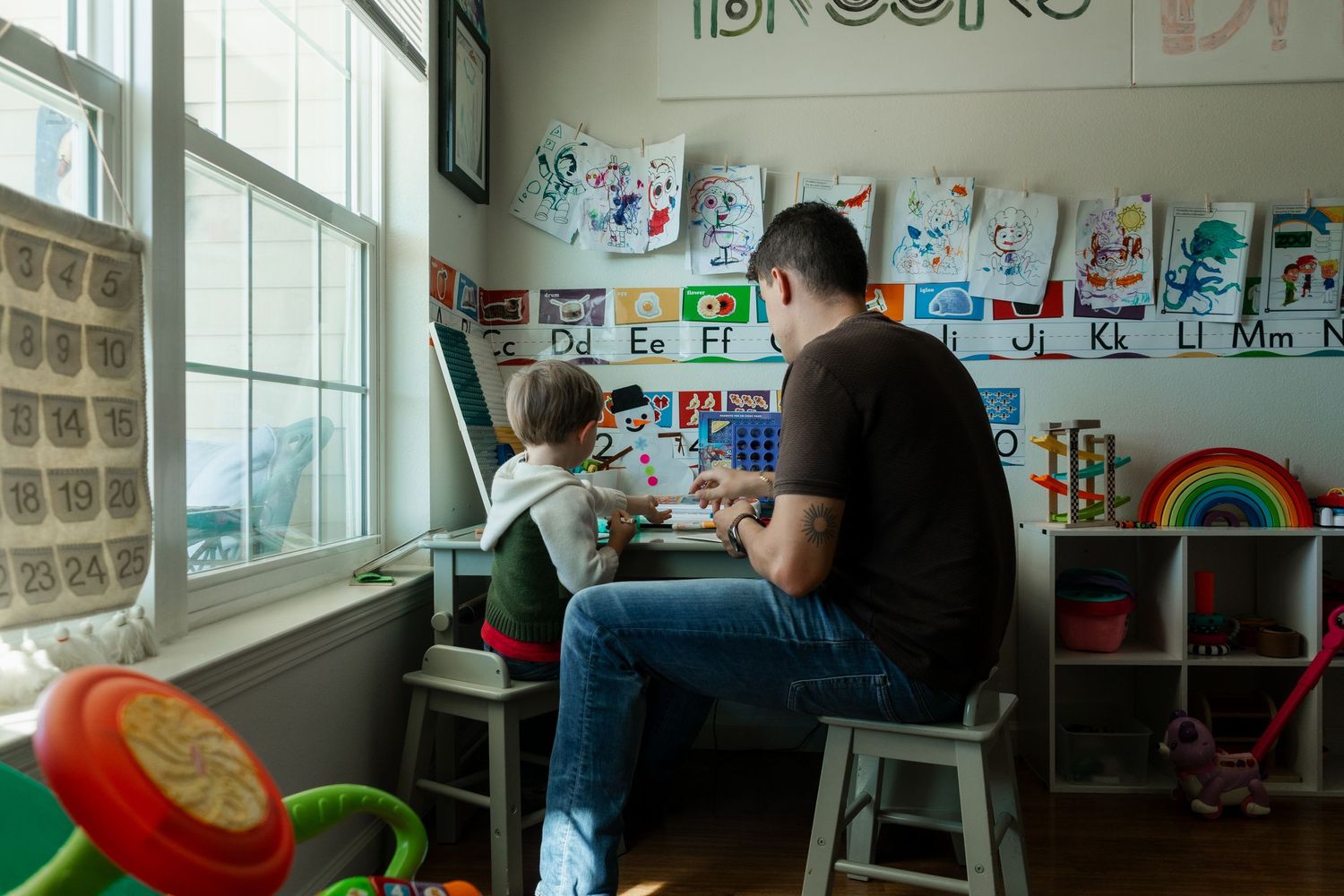 Father and child sitting by window in colorful playroom with children's artwork displayed on walls.