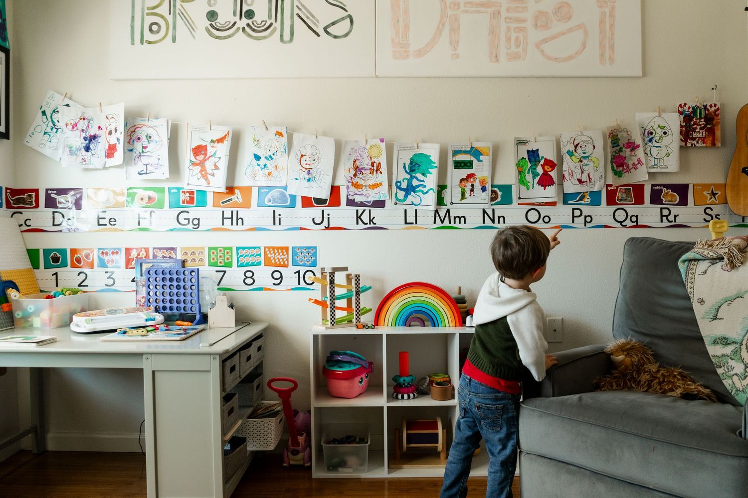 Young child standing in front of colorful alphabet wall display with children's artwork in playroom.