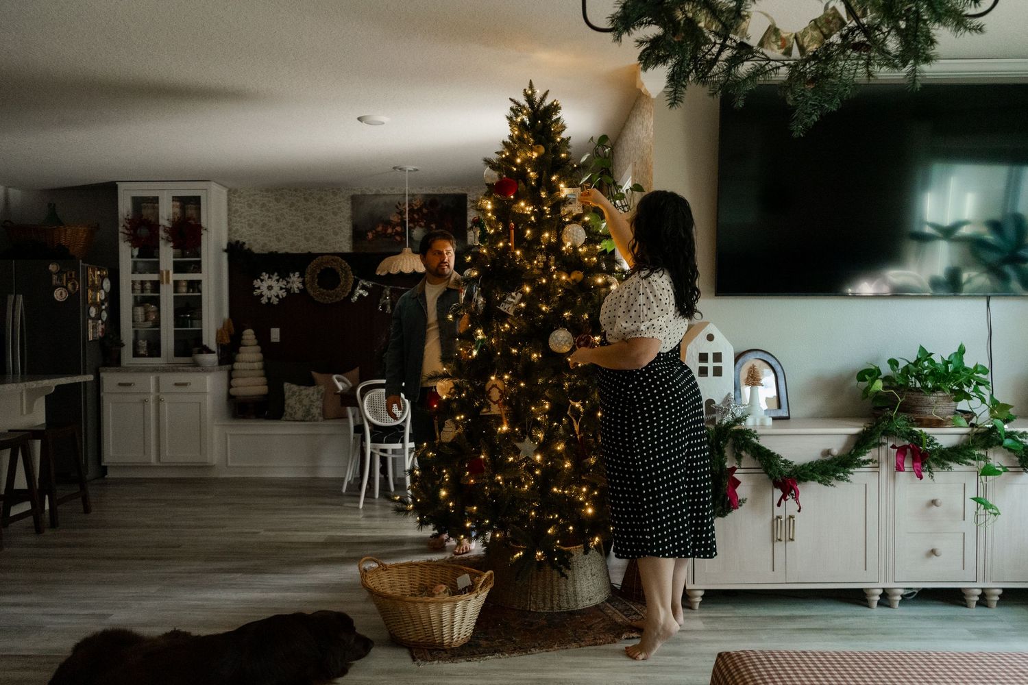 Woman decorating a lit Christmas tree in a festive living room with holiday garlands and warm seasonal decor.