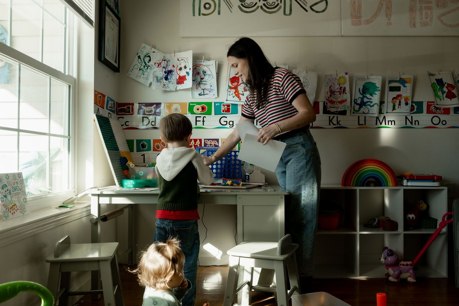 Mother interacts with two young children in colorful playroom with alphabet and artwork wall.