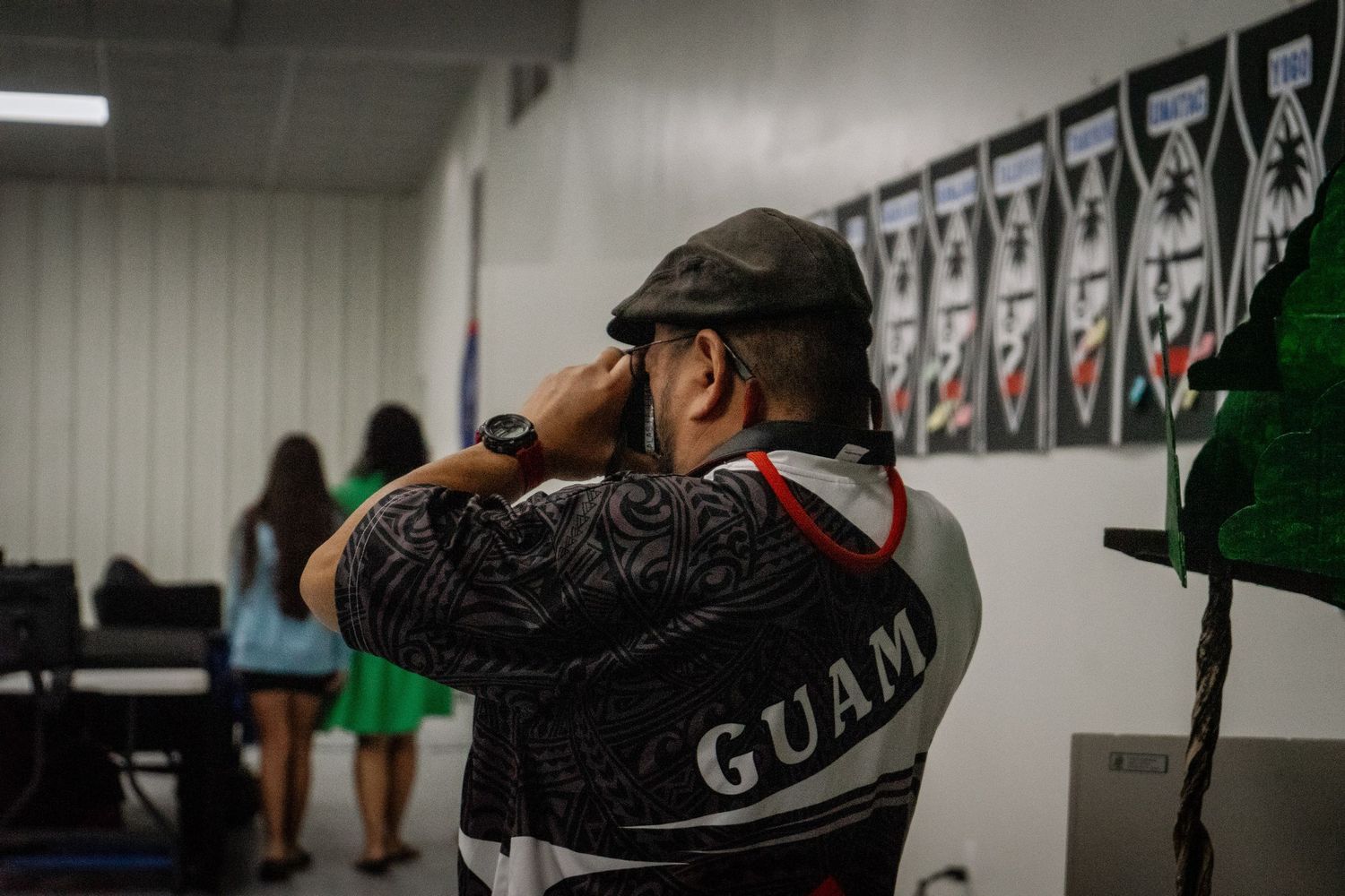 A man wearing a Guam jersey salutes in front of traditional Polynesian artwork displays.