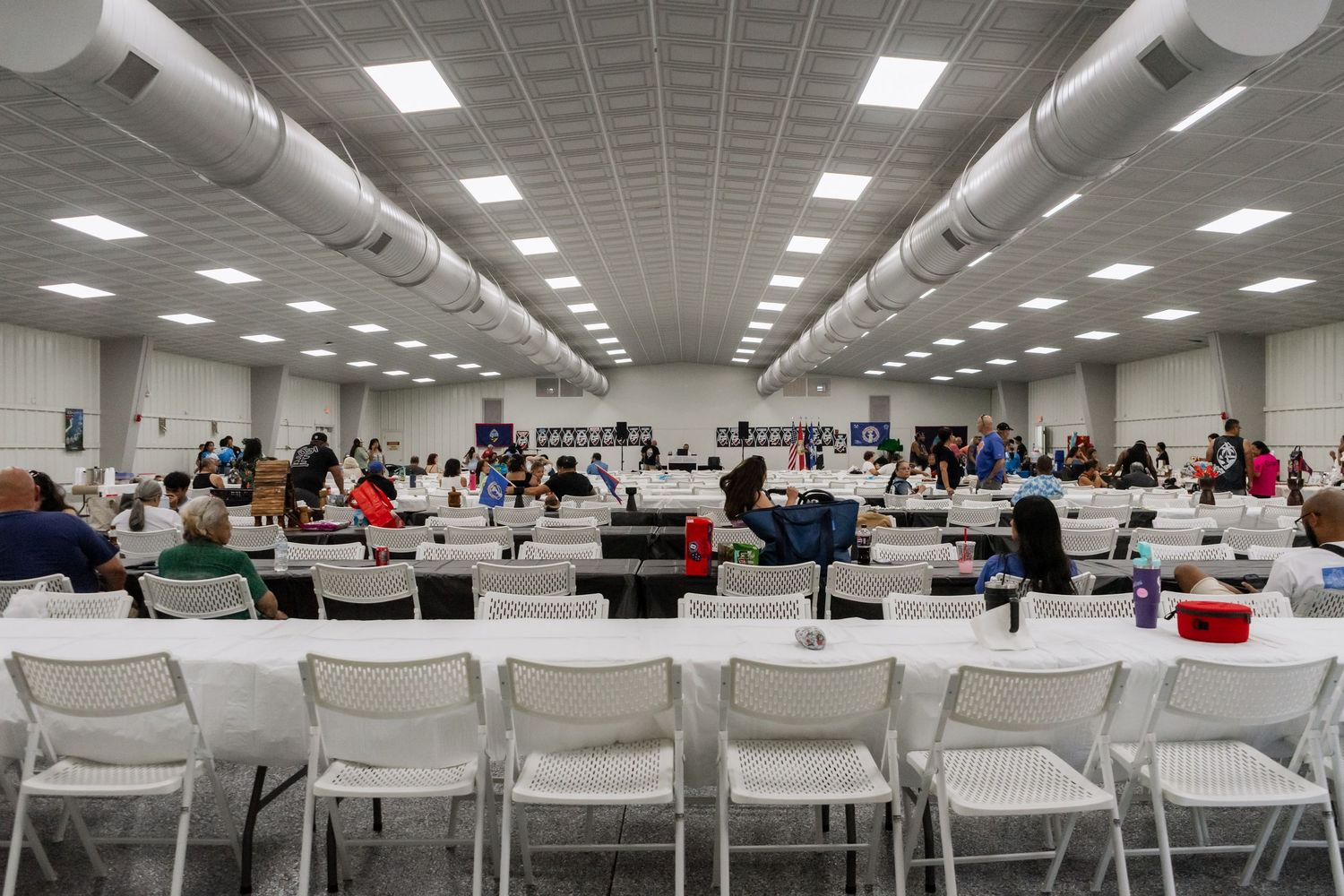 A large indoor community gathering hall filled with people seated at long white tables.