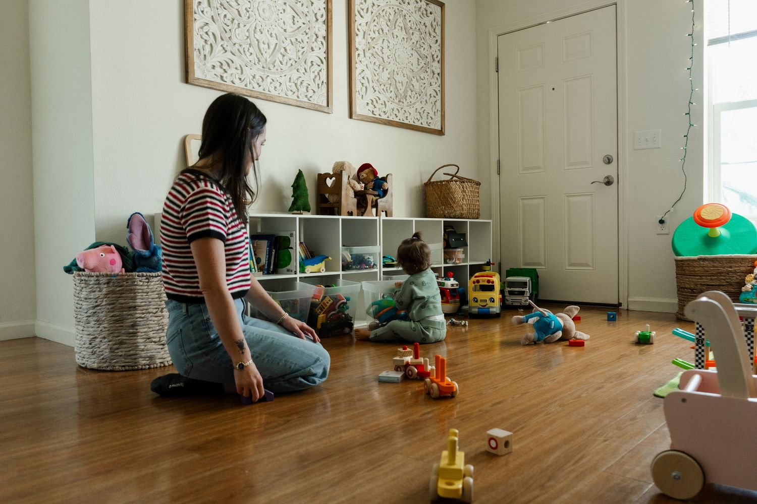 Woman sitting on hardwood floor watching toddler play with colorful toys near white shelving unit in bright playroom.