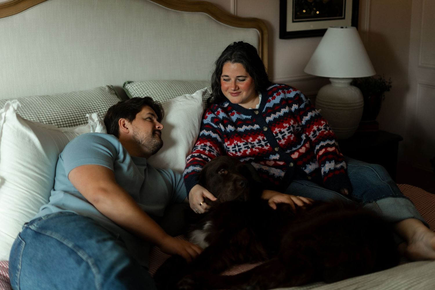 Two people relaxing on a bed with a dark brown dog, in a cozy warmly lit room.