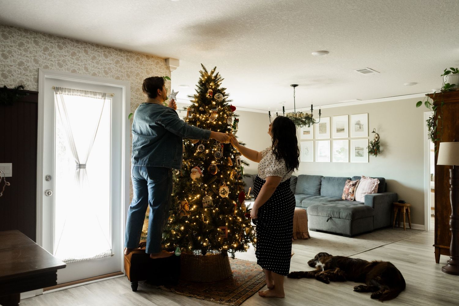 Couple decorating a lit Christmas tree in a cozy living room with a dog resting nearby.