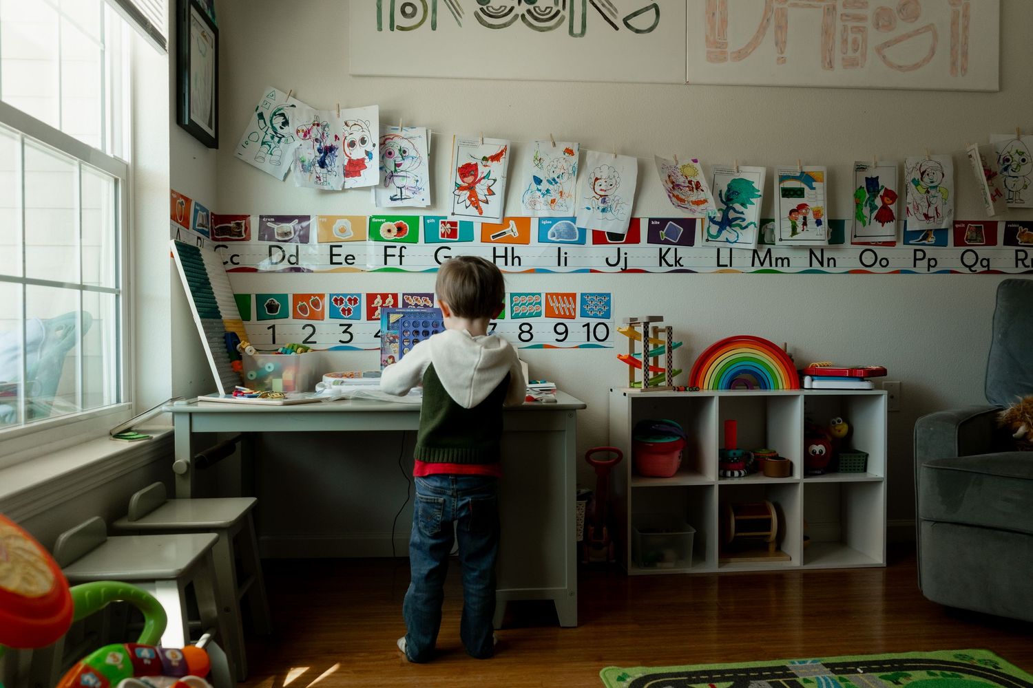Young child stands at a desk in a colorful classroom with alphabet charts and artwork on the walls.