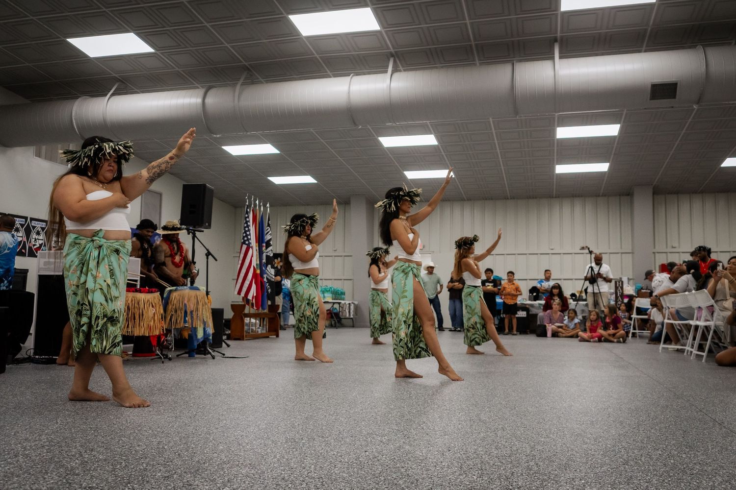 Group of women in grass skirts perform traditional Hawaiian hula dance at indoor cultural festival.