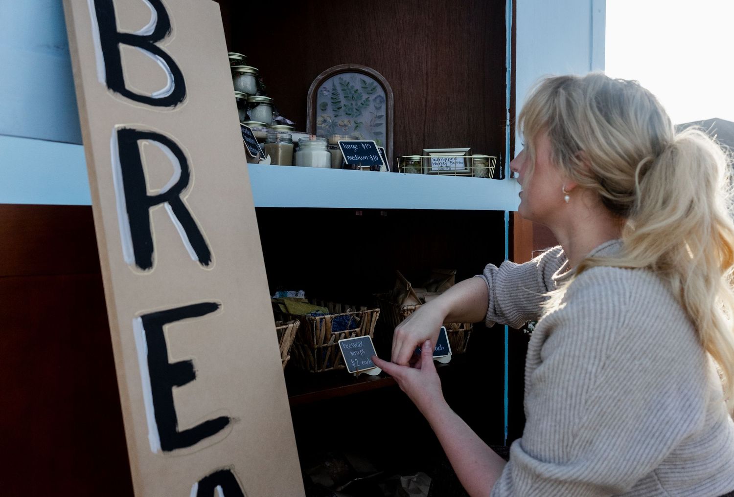 Woman scanning a product at a rustic bread stand with wooden sign and shelves of goods.