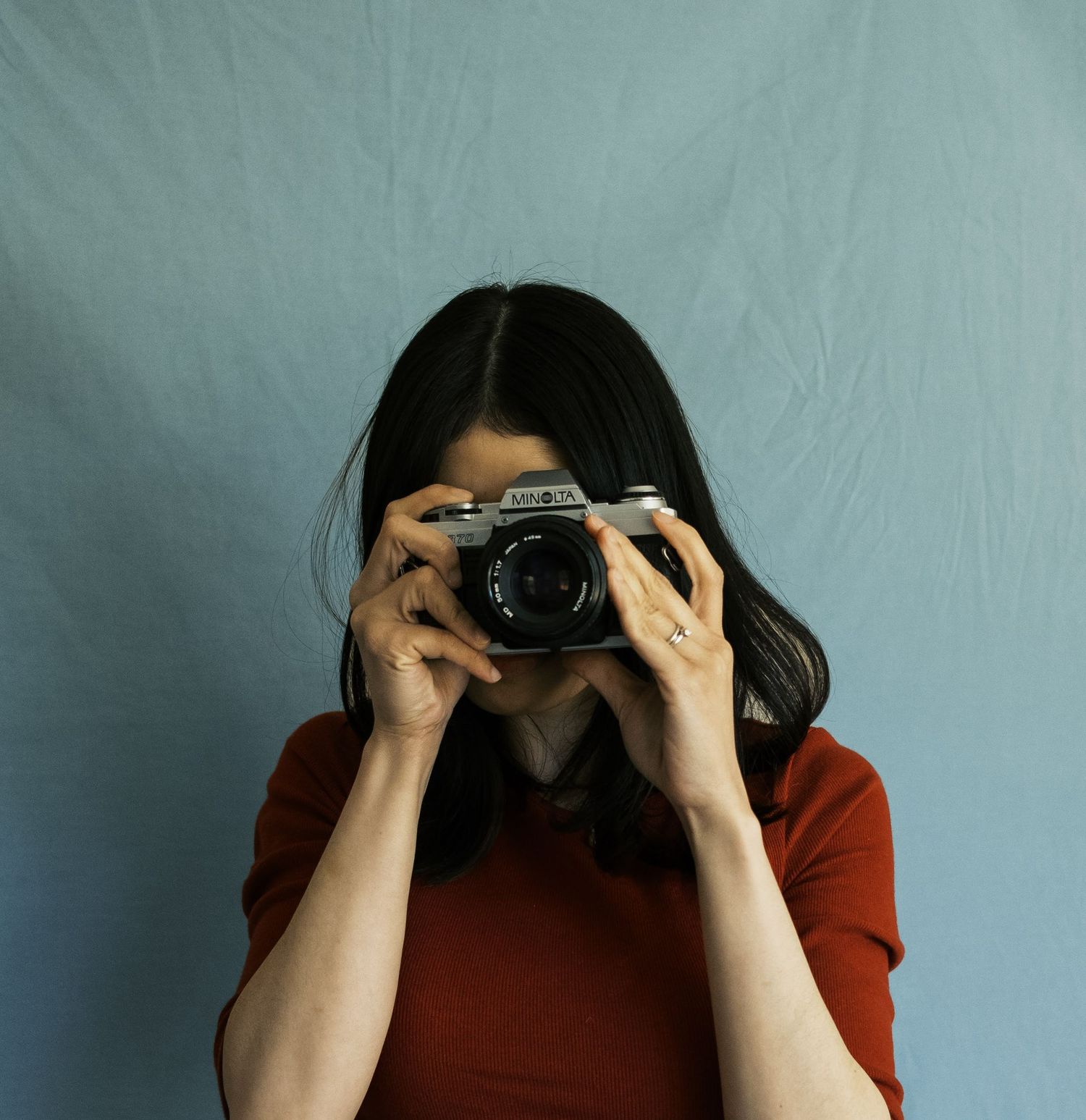 Woman in a red top holds a vintage Minolta film camera up to her face against a light blue backdrop.