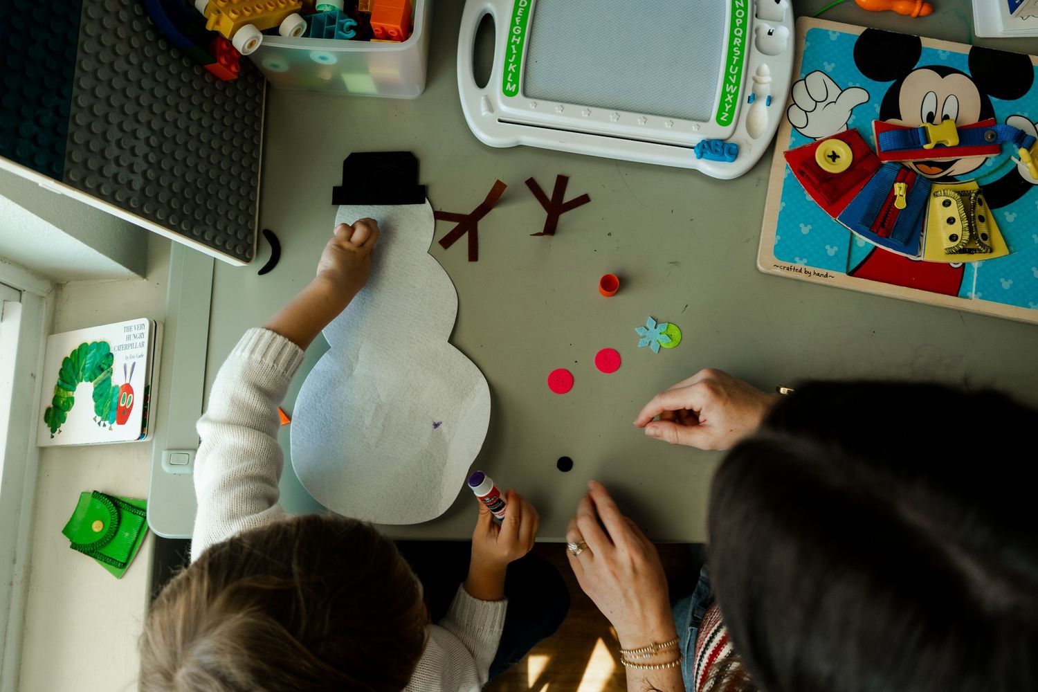 Overhead view of child and adult creating a snowman craft with felt pieces on a table.