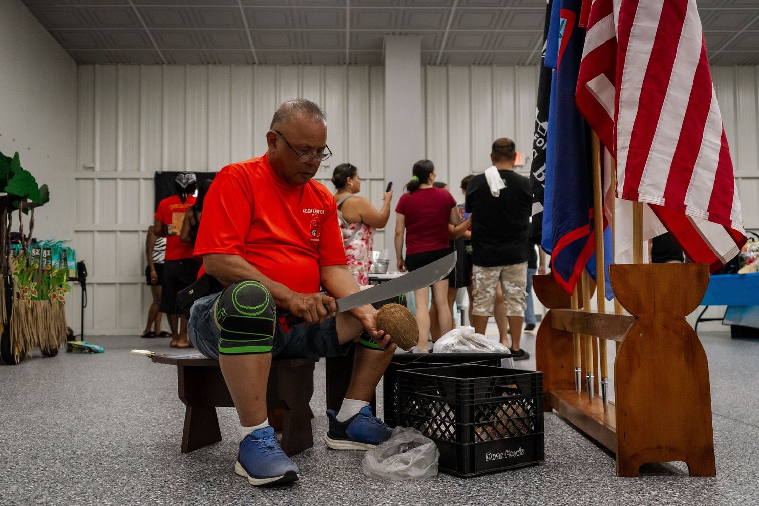 Man in red shirt sits on bench near American flag preparing for cultural performance at indoor event.