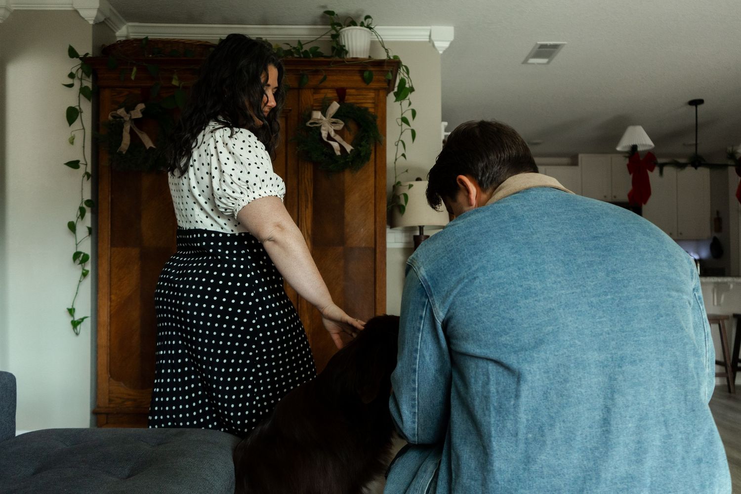 A woman in a polka dot skirt stands near a wooden cabinet while a man sits on a couch in a cozy living room.