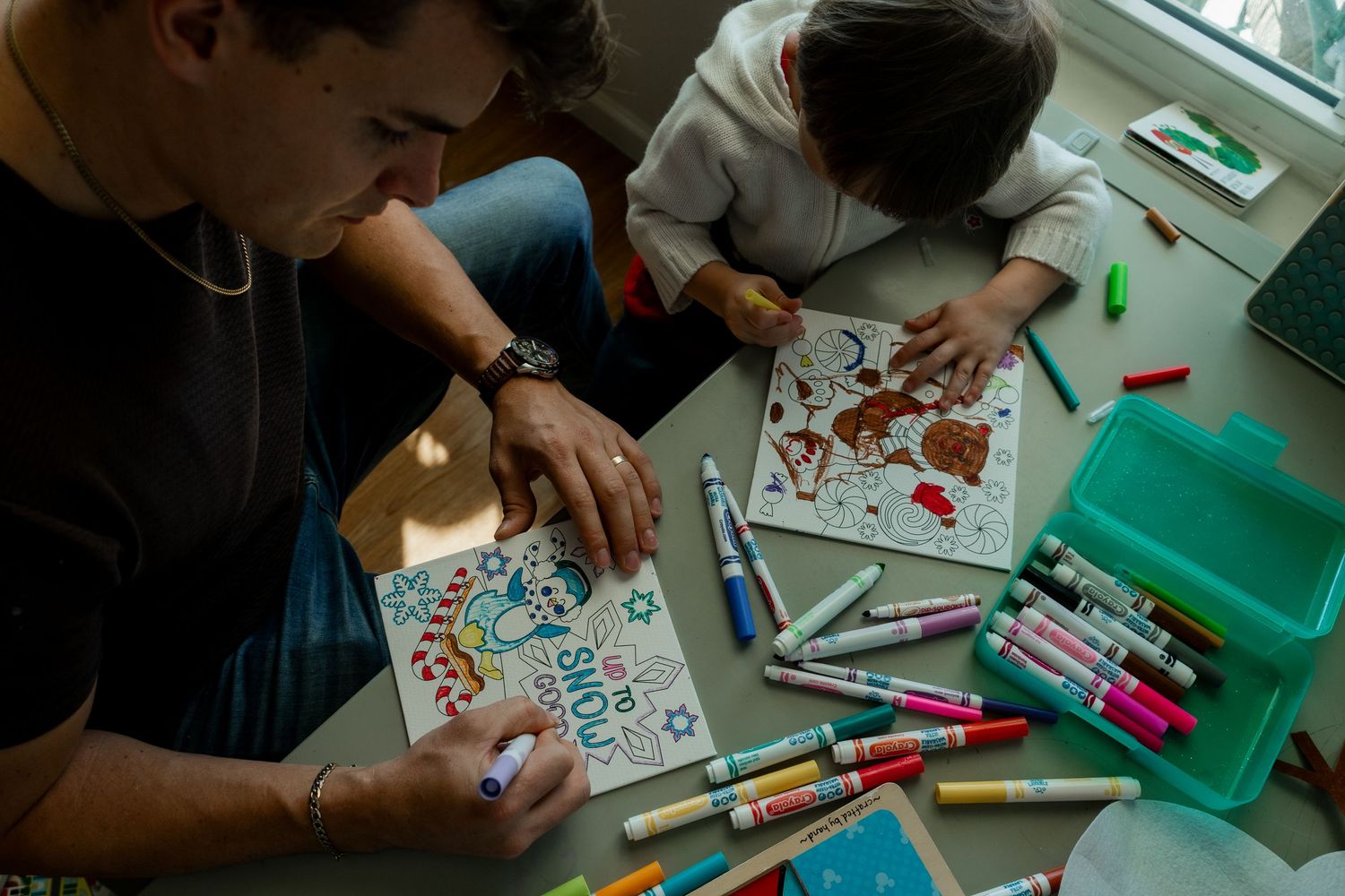 Adults and child coloring together at a table with markers and coloring books in warm indoor light.