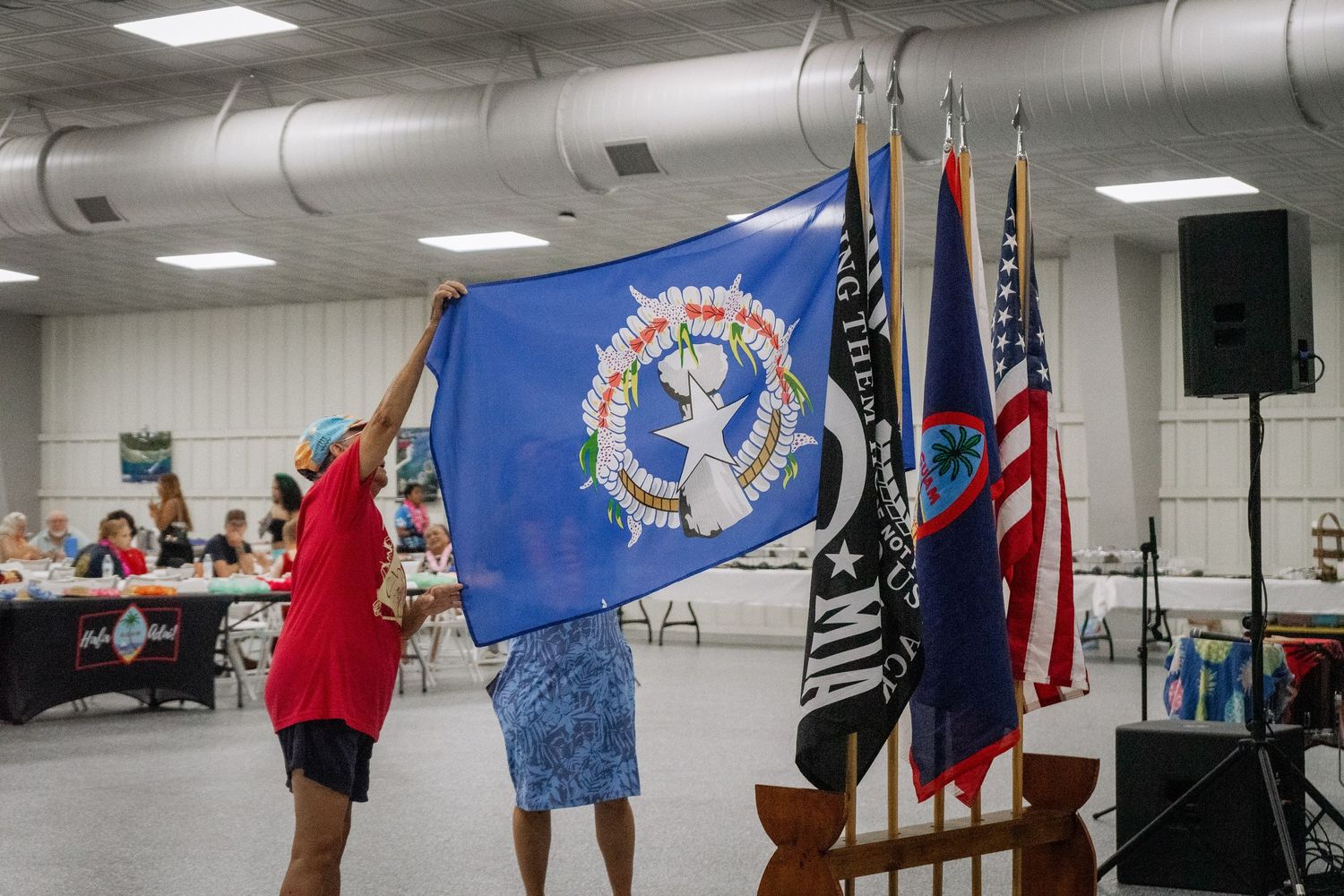 Two people display the Northern Mariana Islands flag at an indoor cultural ceremony.