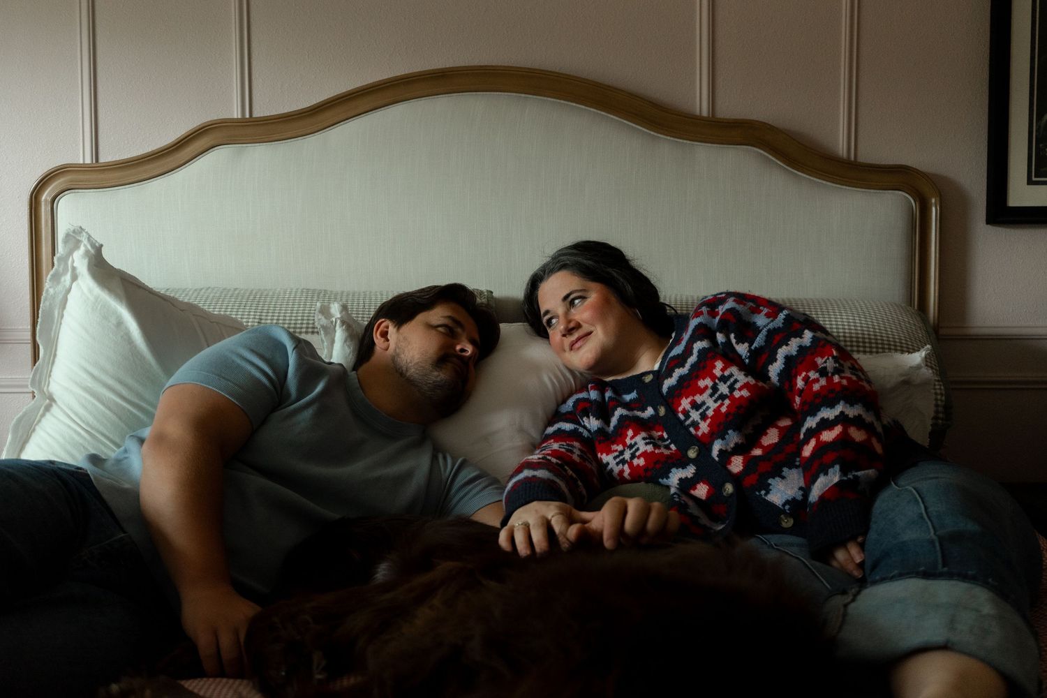 Couple lying on a bed smiling at each other, with a dog resting beside them in a cozy bedroom.