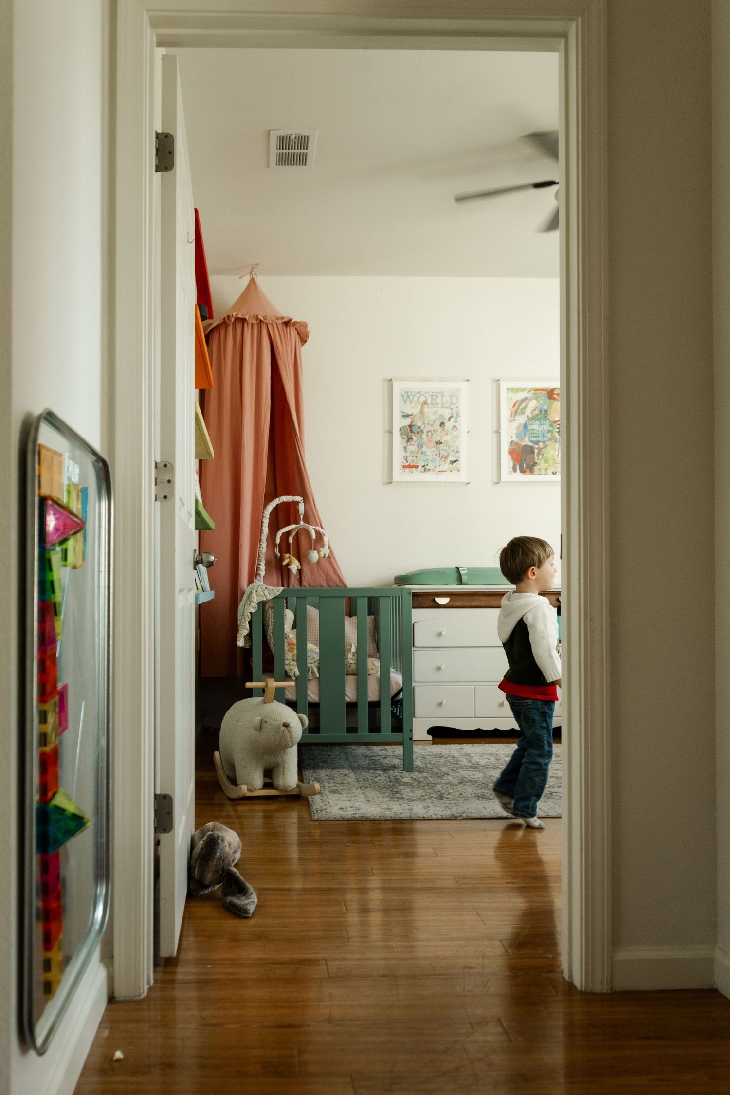 Young child stands in nursery doorway with green crib, canopy, and stuffed animals visible.