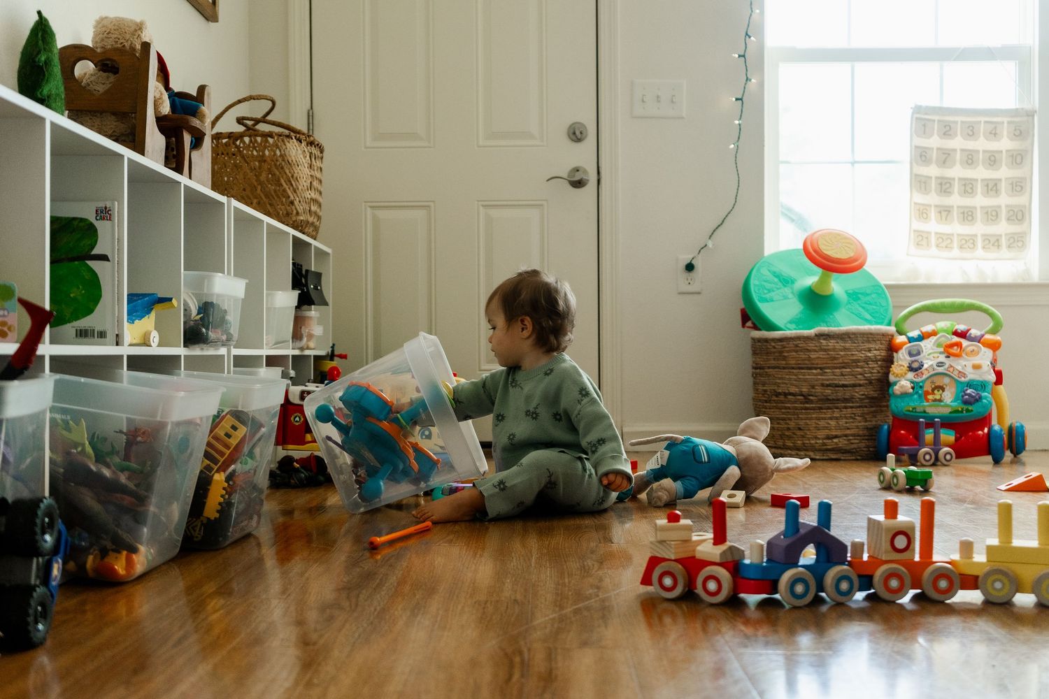 Toddler in green outfit sitting on hardwood floor playing with wooden train toys beside white storage shelves.
