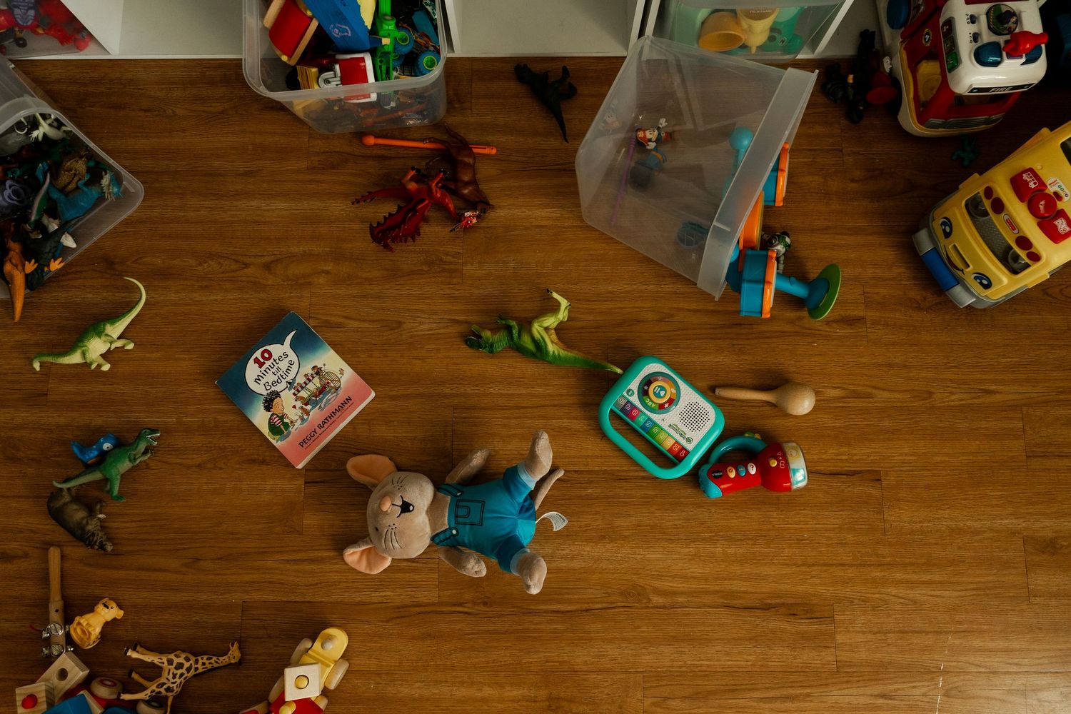 Collection of children's toys scattered on hardwood floor including dinosaurs and stuffed animals.