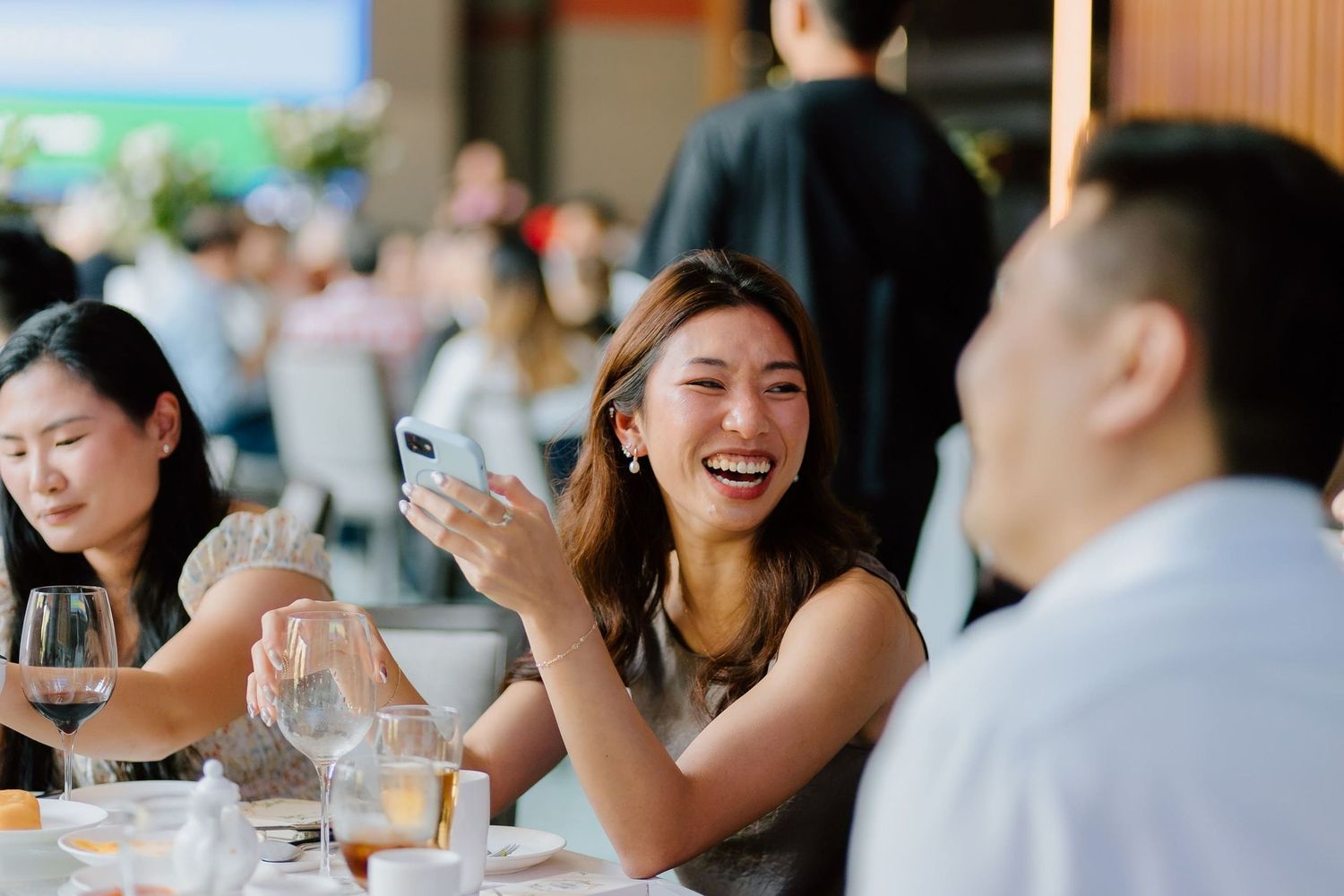 A smiling woman holding a phone laughs with guests at an elegant wedding reception dinner table.