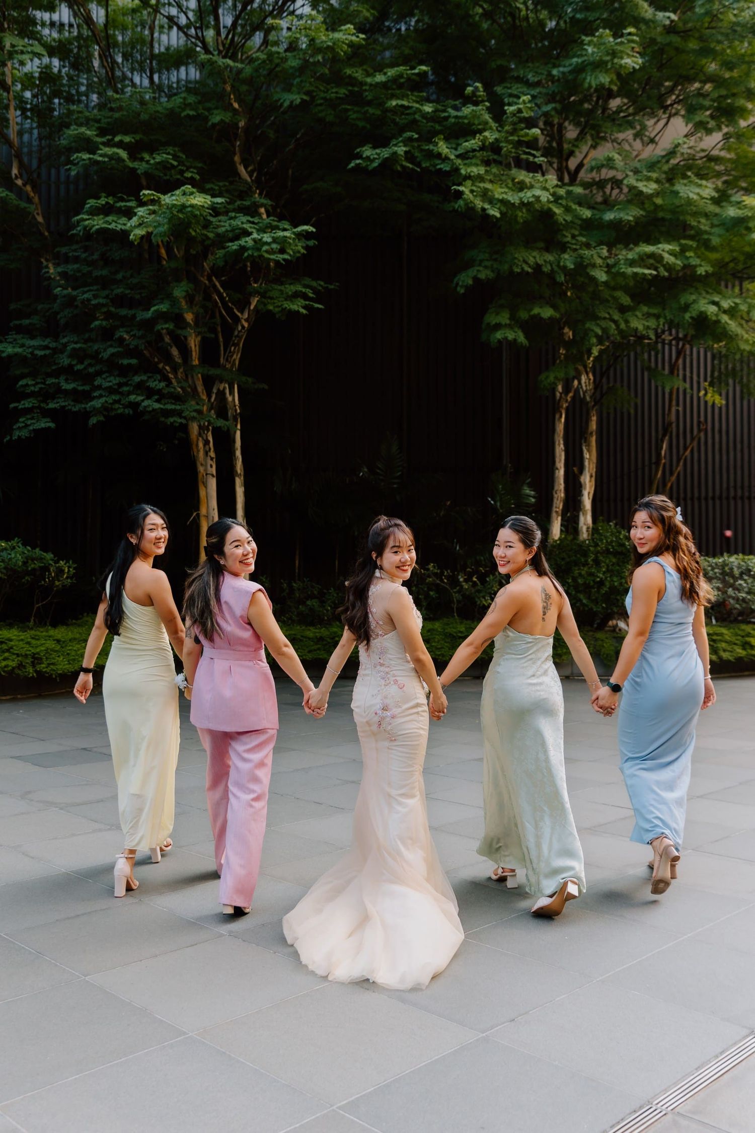 Bride in white gown holds hands with four bridesmaids in pastel dresses, walking outdoors near lush green trees.