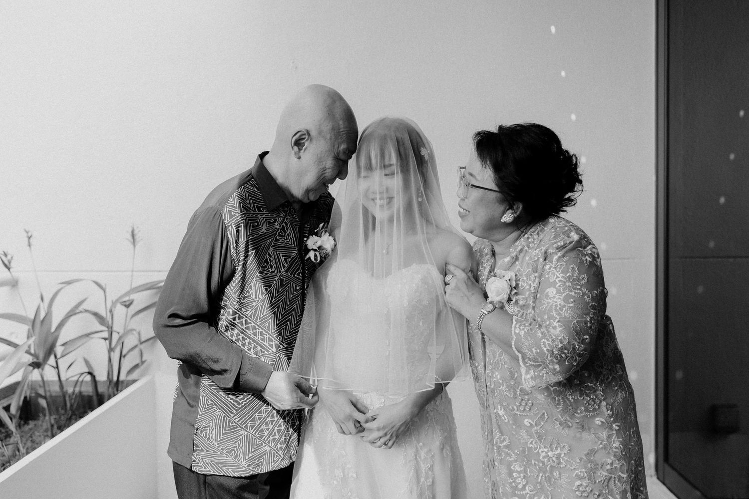 Bride in white gown and veil shares tender moment with parents on wedding day in black and white photo.