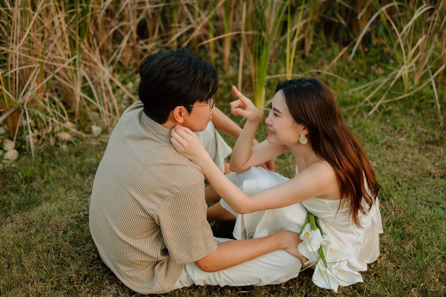 Couple sitting on grass near tall reeds, woman touching man's face and smiling intimately outdoors.