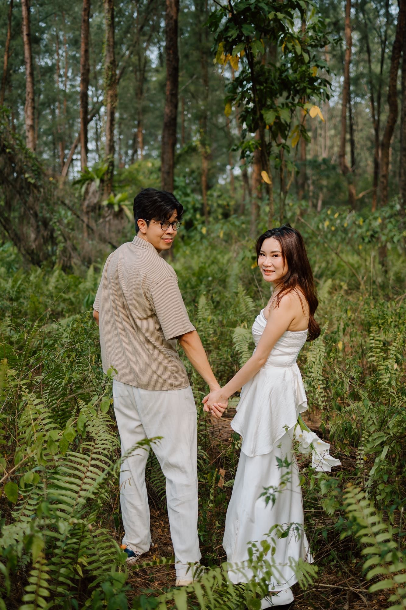 Couple holding hands in a lush forest, woman in white dress, man in beige shirt and white pants.